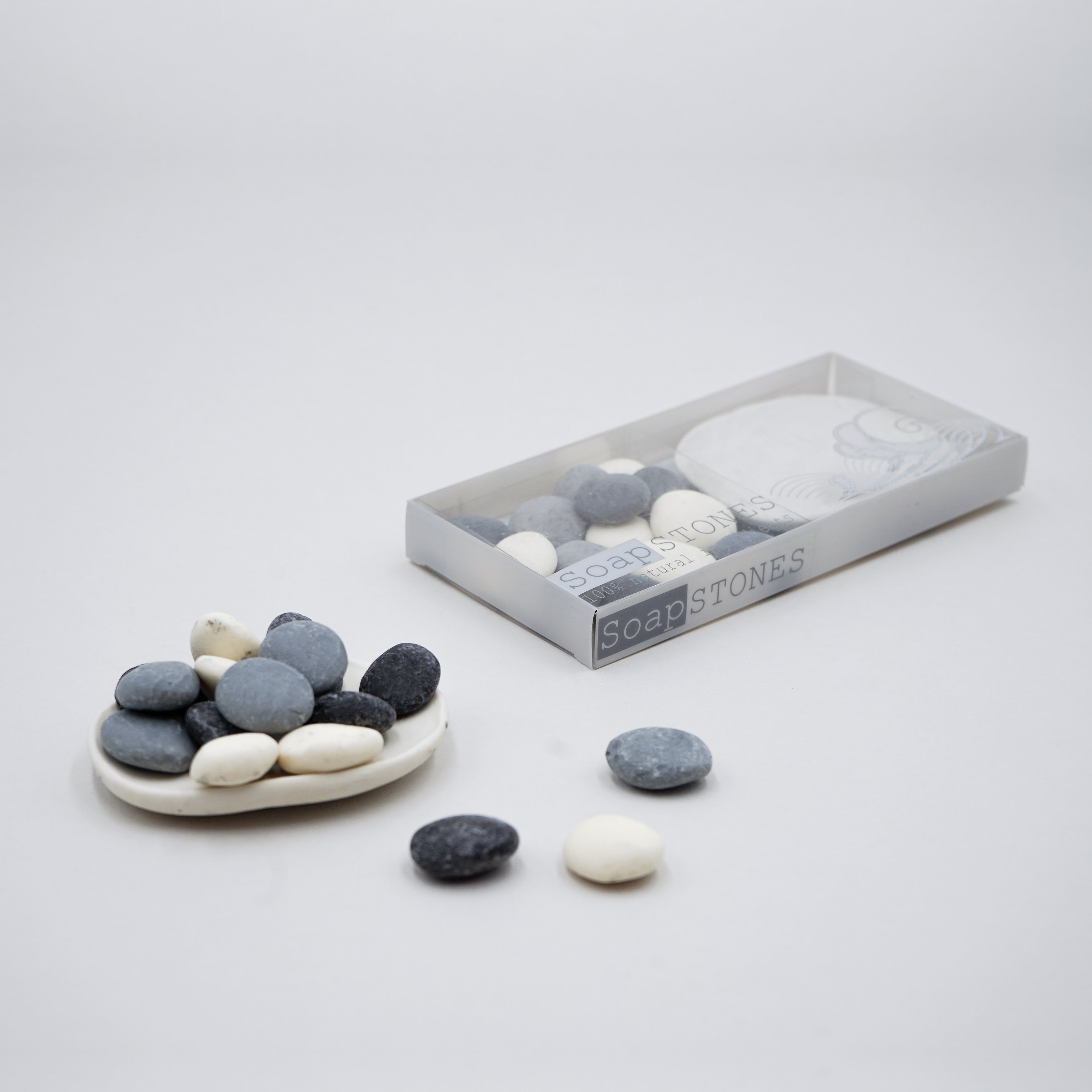 A set of assorted small pebble soaps in various colors displayed on a white porcelain dish with a grey marble effect.