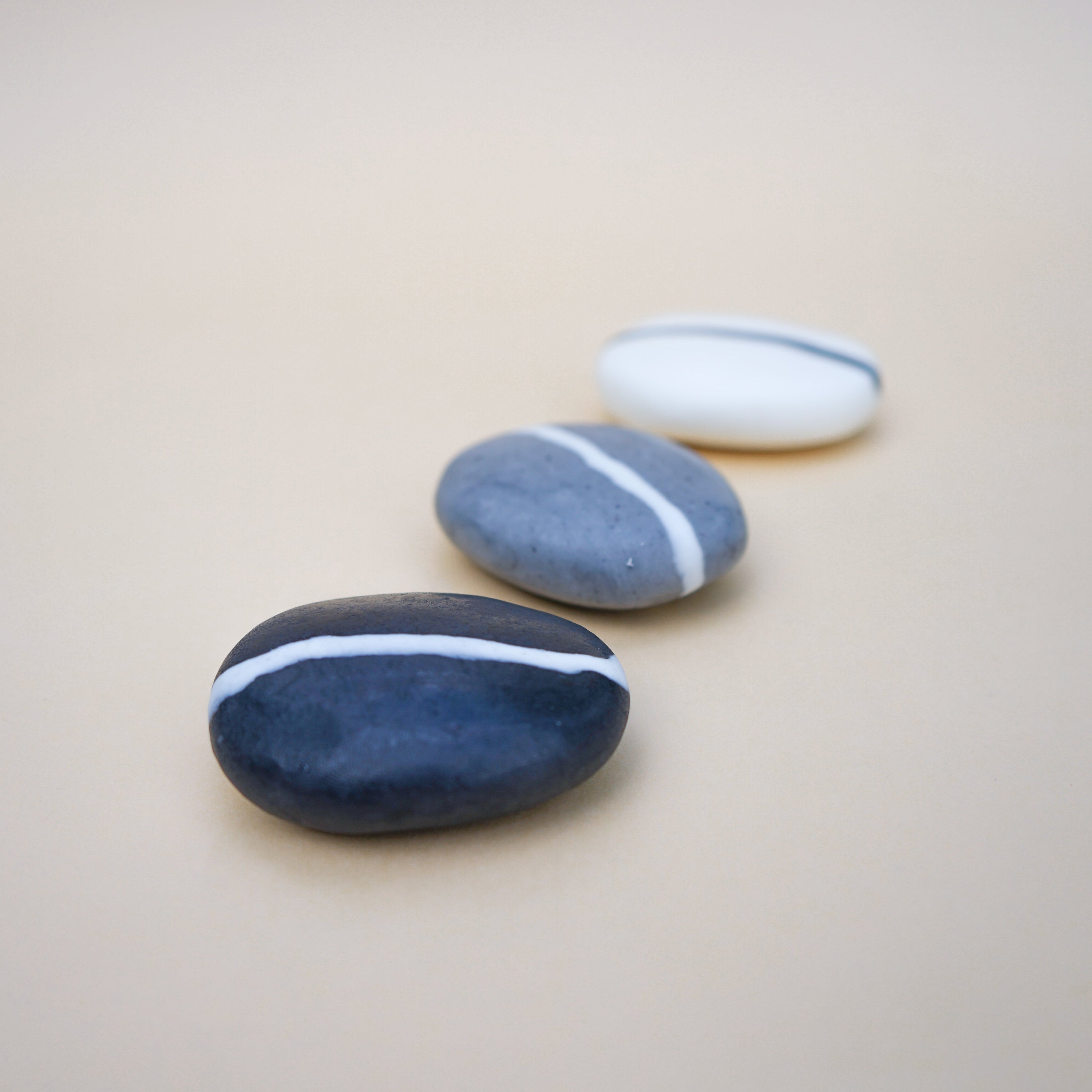 A set of three handmade pebble-shaped soaps in charcoal, grey, and white with a stylish stripe design, showcasing their natural beauty.