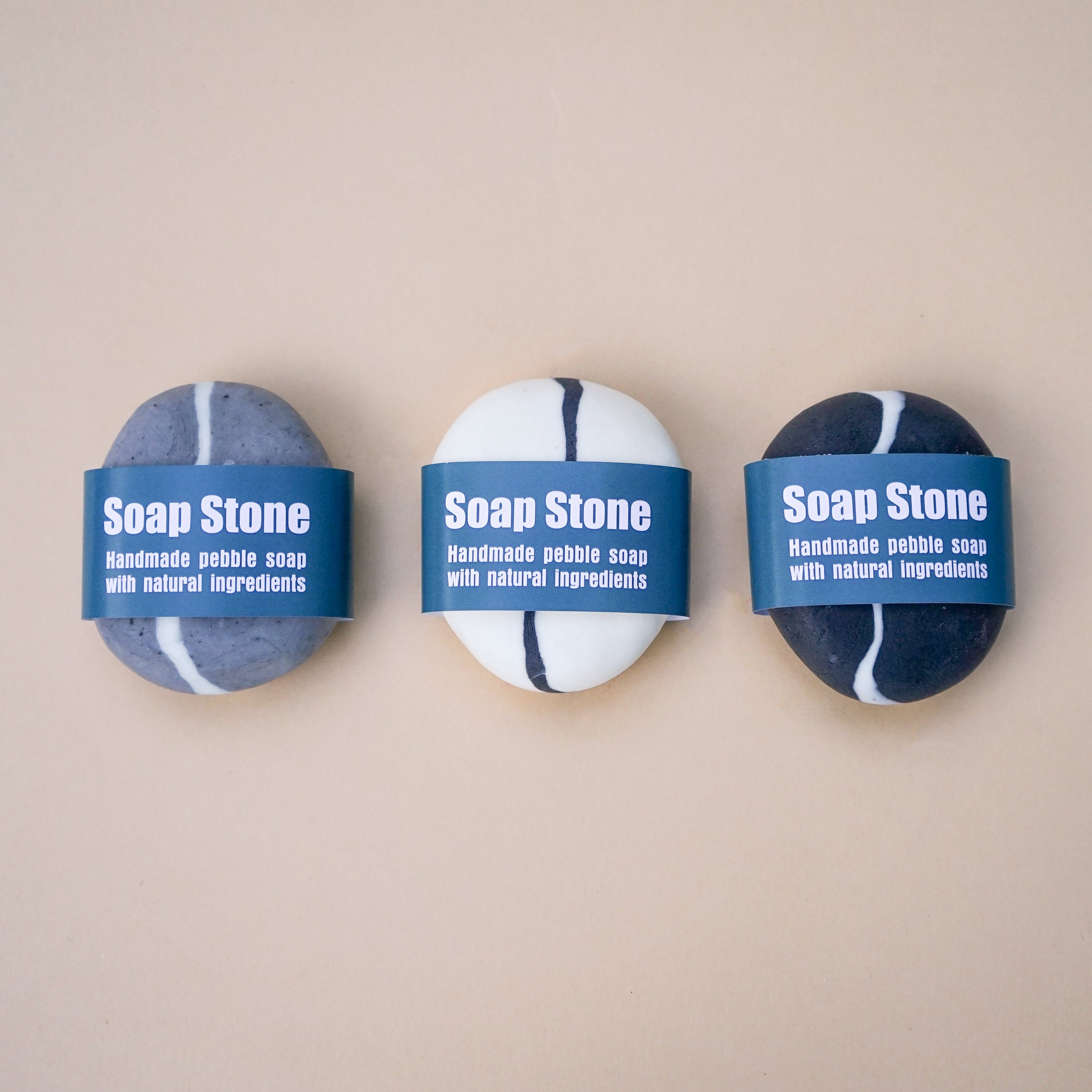 A set of three handmade pebble-shaped soaps in charcoal, grey, and white with a stylish stripe design, showcasing their natural beauty.