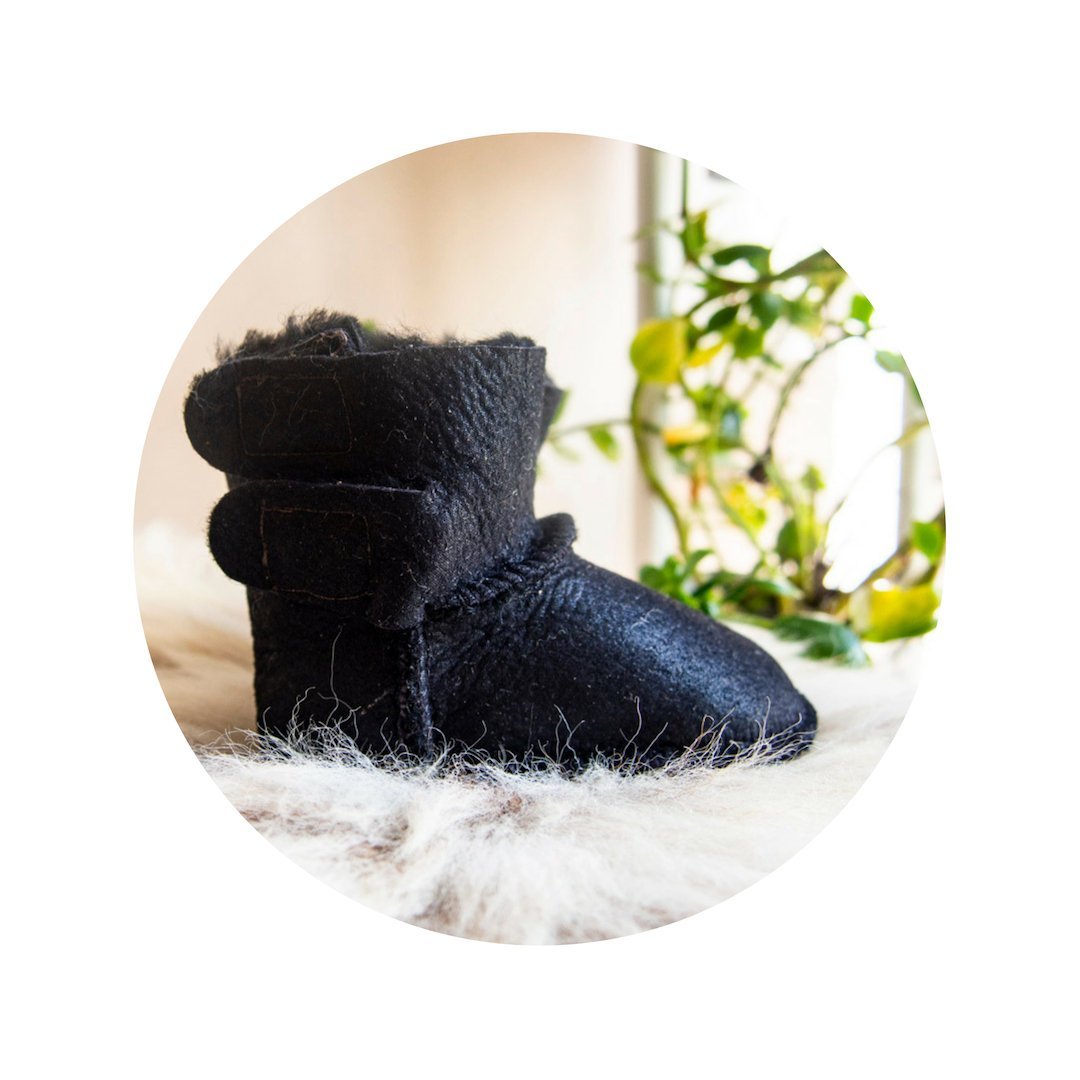 A pair of handmade baby black sheepskin boots with soft fur lining, featuring velcro fastenings for easy wear.