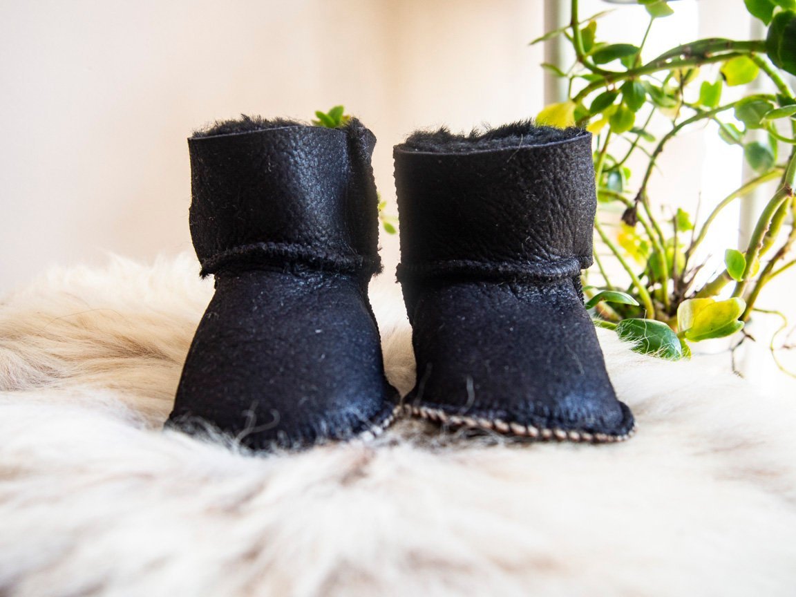 A pair of handmade baby black sheepskin boots with soft fur lining, featuring velcro fastenings for easy wear.