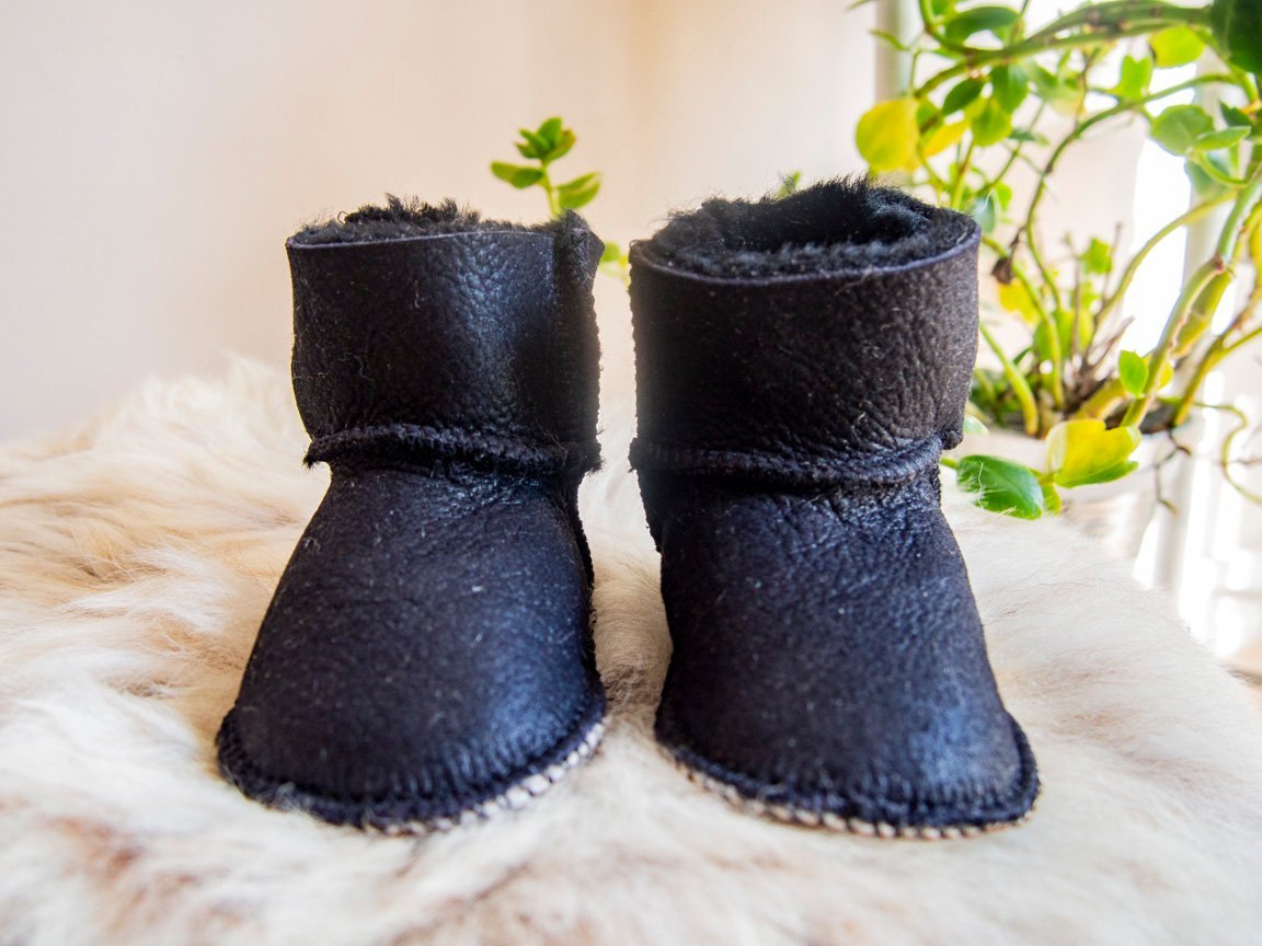 A pair of handmade baby black sheepskin boots with soft fur lining, featuring velcro fastenings for easy wear.