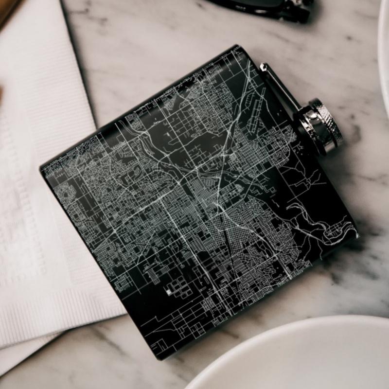 Matte black hip flask featuring a custom engraved map of Bakersfield, California, with coordinates, accompanied by a canvas bag and funnel.