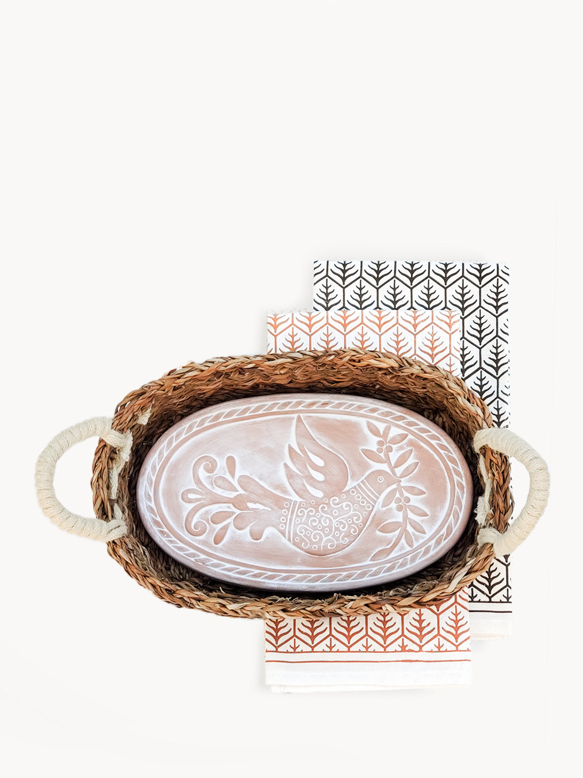 A beautifully crafted terracotta bread warmer with a bird design, accompanied by a handwoven seagrass basket and a natural cotton tea towel.