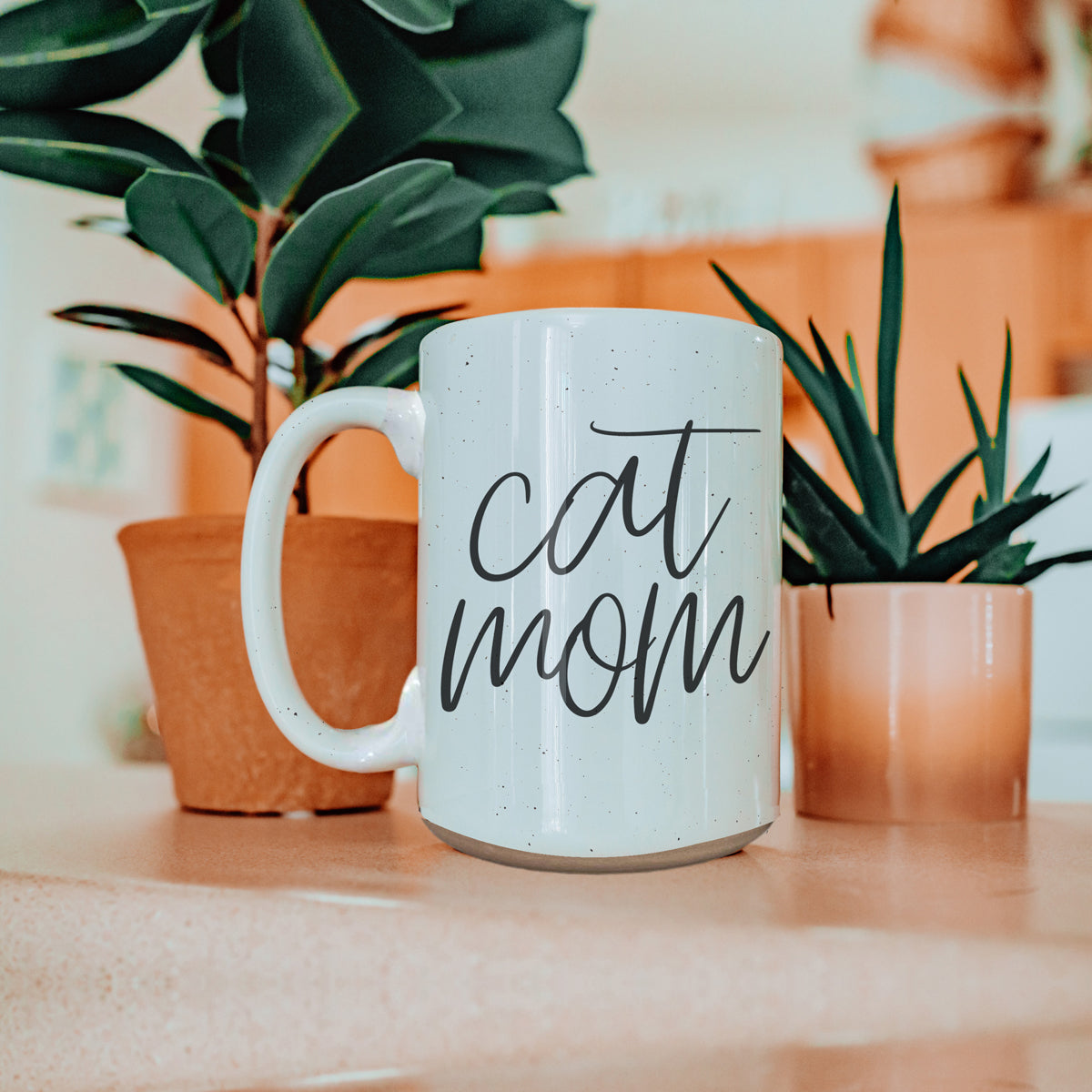 A stylish off-white ceramic mug with dark speckles, featuring a black graphic that reads 'Cat Mom' on both sides, perfect for cat lovers.