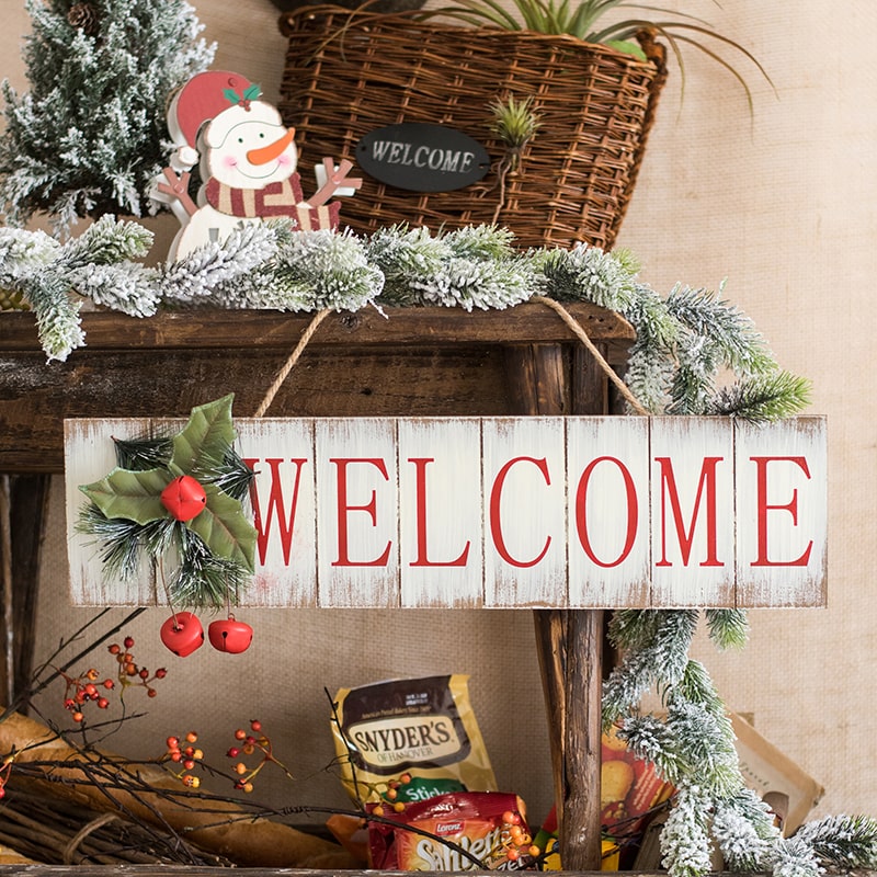 A festive Christmas Welcome Sign made of wood, featuring vibrant red, white, and green colors, adorned with faux plants and a hemp rope for hanging.