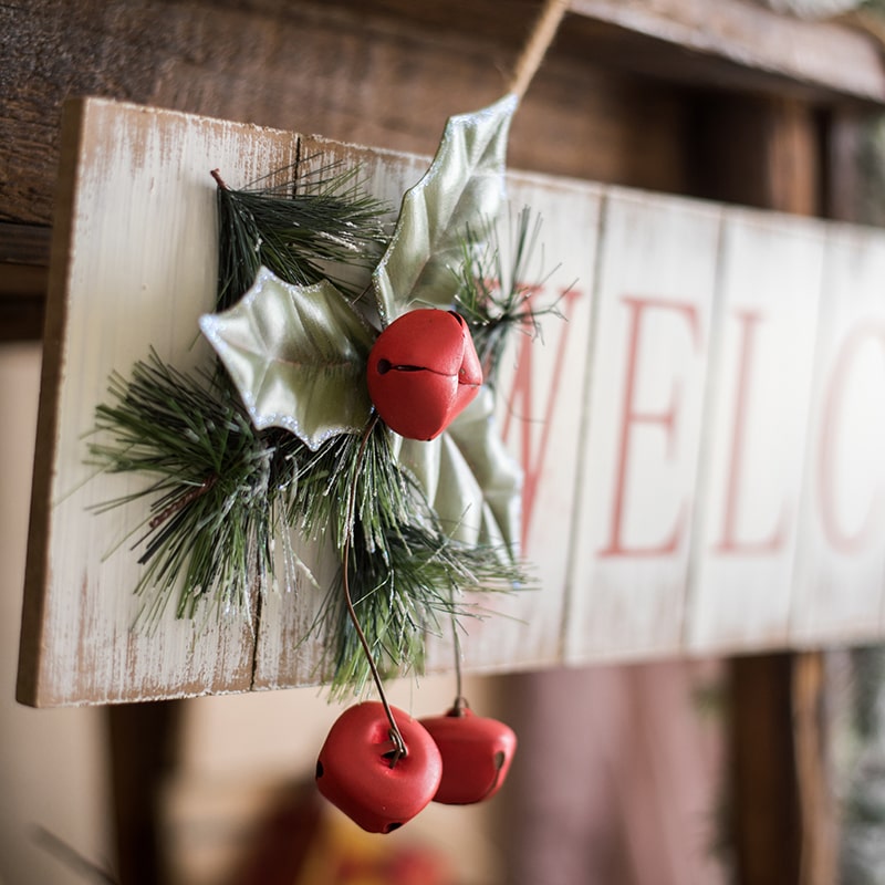 A festive Christmas Welcome Sign made of wood, featuring vibrant red, white, and green colors, adorned with faux plants and a hemp rope for hanging.