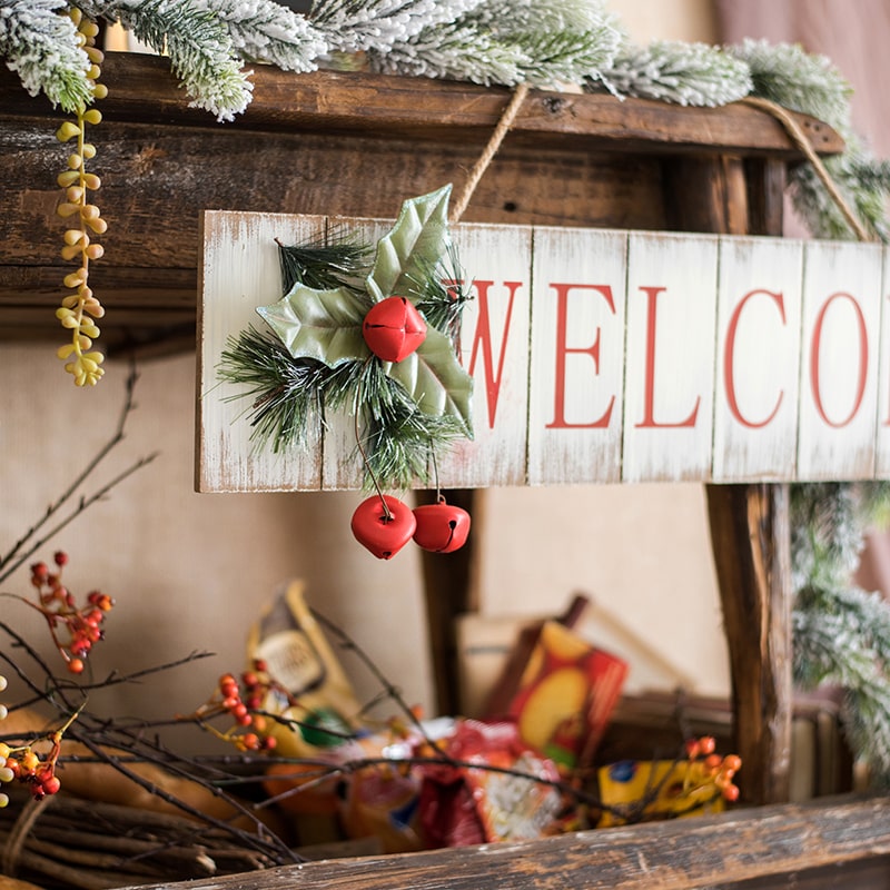 A festive Christmas Welcome Sign made of wood, featuring vibrant red, white, and green colors, adorned with faux plants and a hemp rope for hanging.