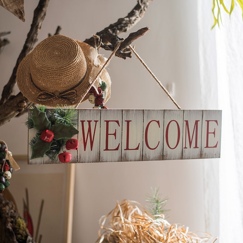 A festive Christmas Welcome Sign made of wood, featuring vibrant red, white, and green colors, adorned with faux plants and a hemp rope for hanging.