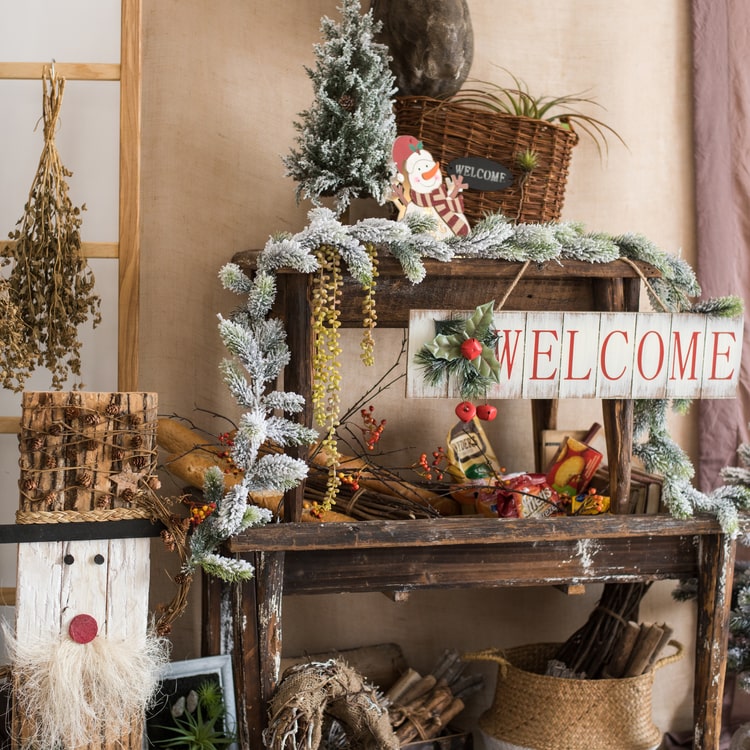 A festive Christmas Welcome Sign made of wood, featuring vibrant red, white, and green colors, adorned with faux plants and a hemp rope for hanging.