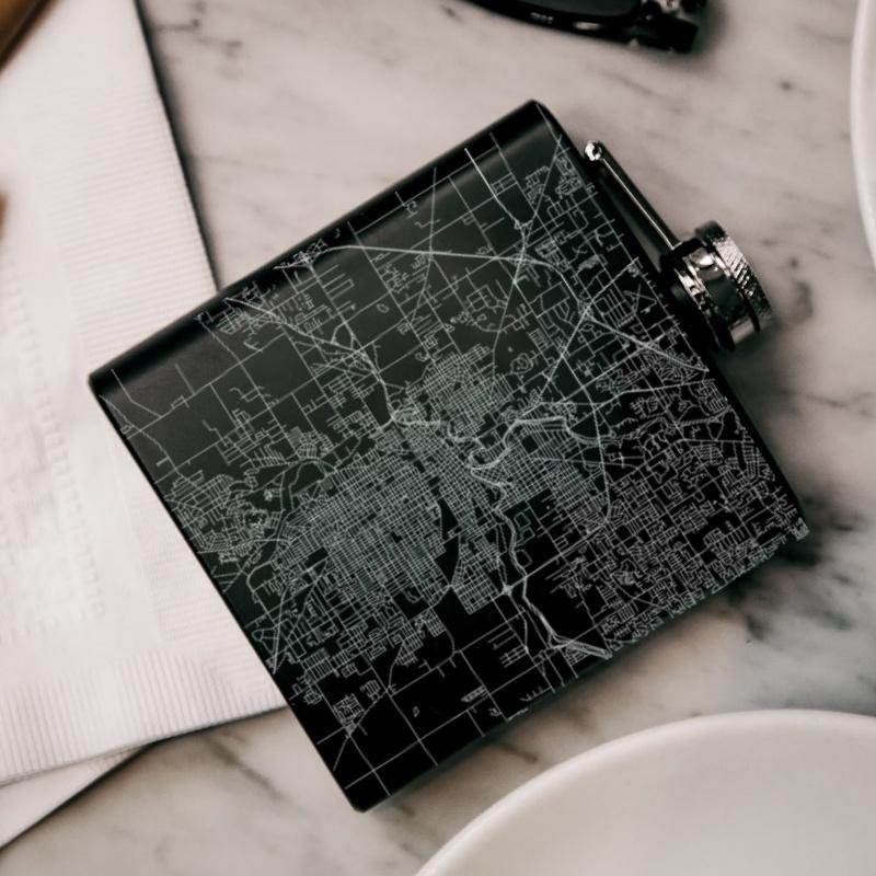 Matte black hip flask featuring a custom engraved map of Fort Wayne, Indiana with coordinates, accompanied by a canvas bag and funnel.