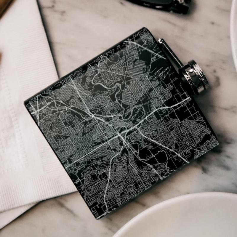 Matte black hip flask featuring a custom engraved map of Fort Worth, Texas, with coordinates, alongside a canvas bag and funnel.