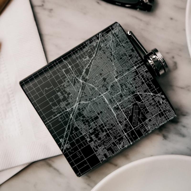 Matte black hip flask featuring a custom engraved map of Fresno, California with coordinates, accompanied by a canvas bag and funnel.