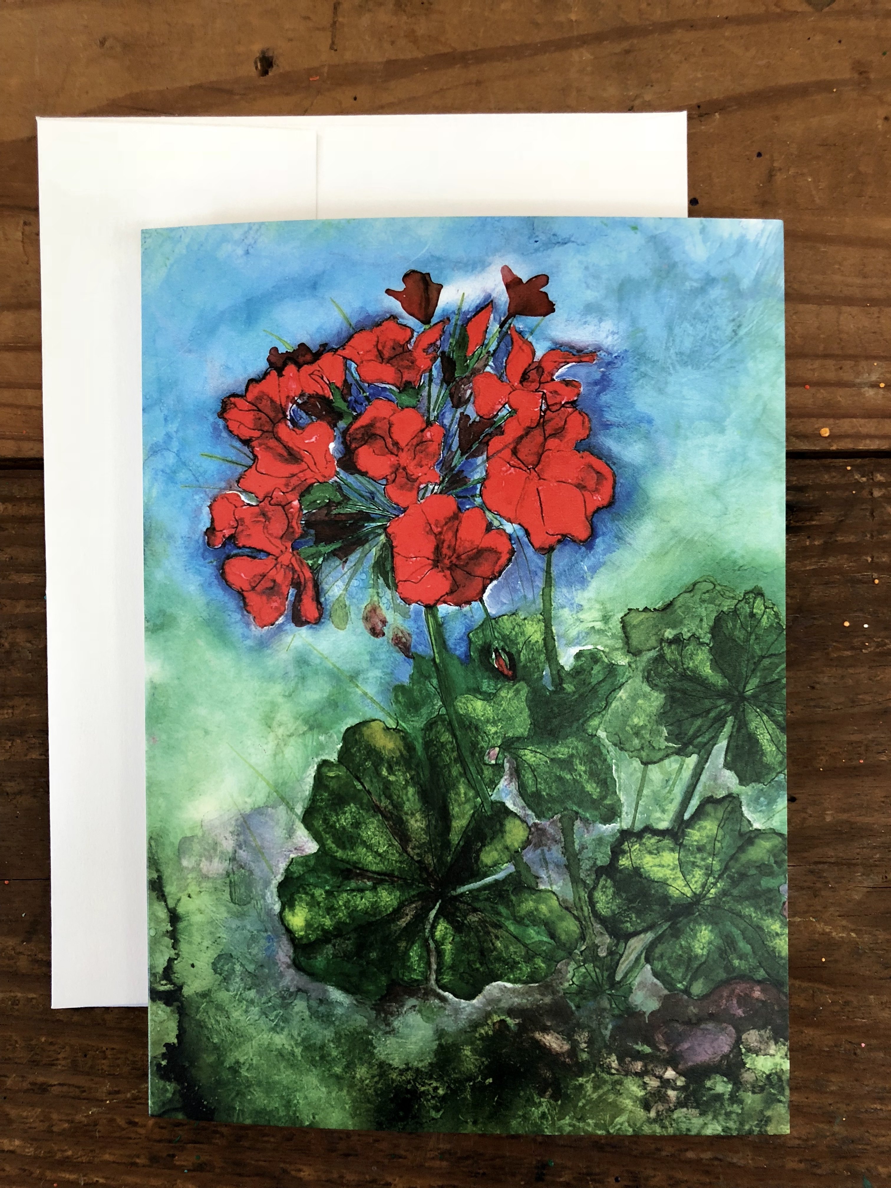 A vibrant red geranium greeting card featuring a mixed media painting with pen, oil paints, and alcohol inks, displayed with a white envelope.