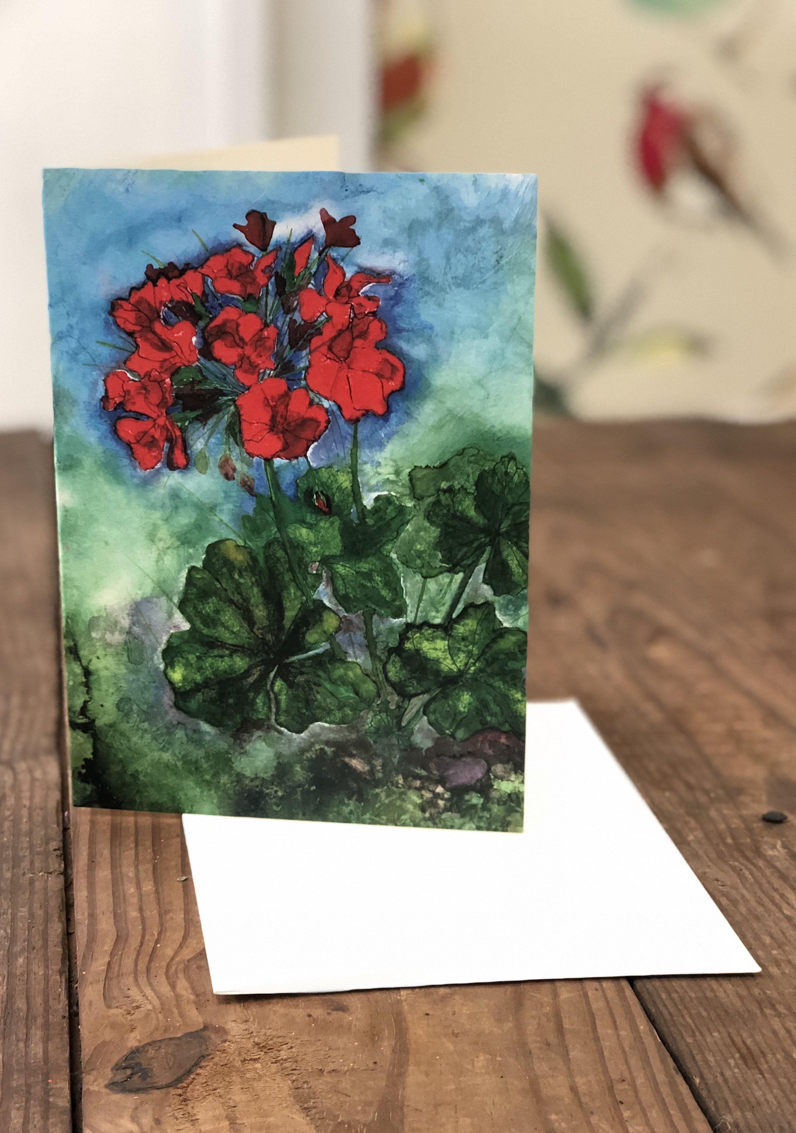 A vibrant red geranium greeting card featuring a mixed media painting with pen, oil paints, and alcohol inks, displayed with a white envelope.