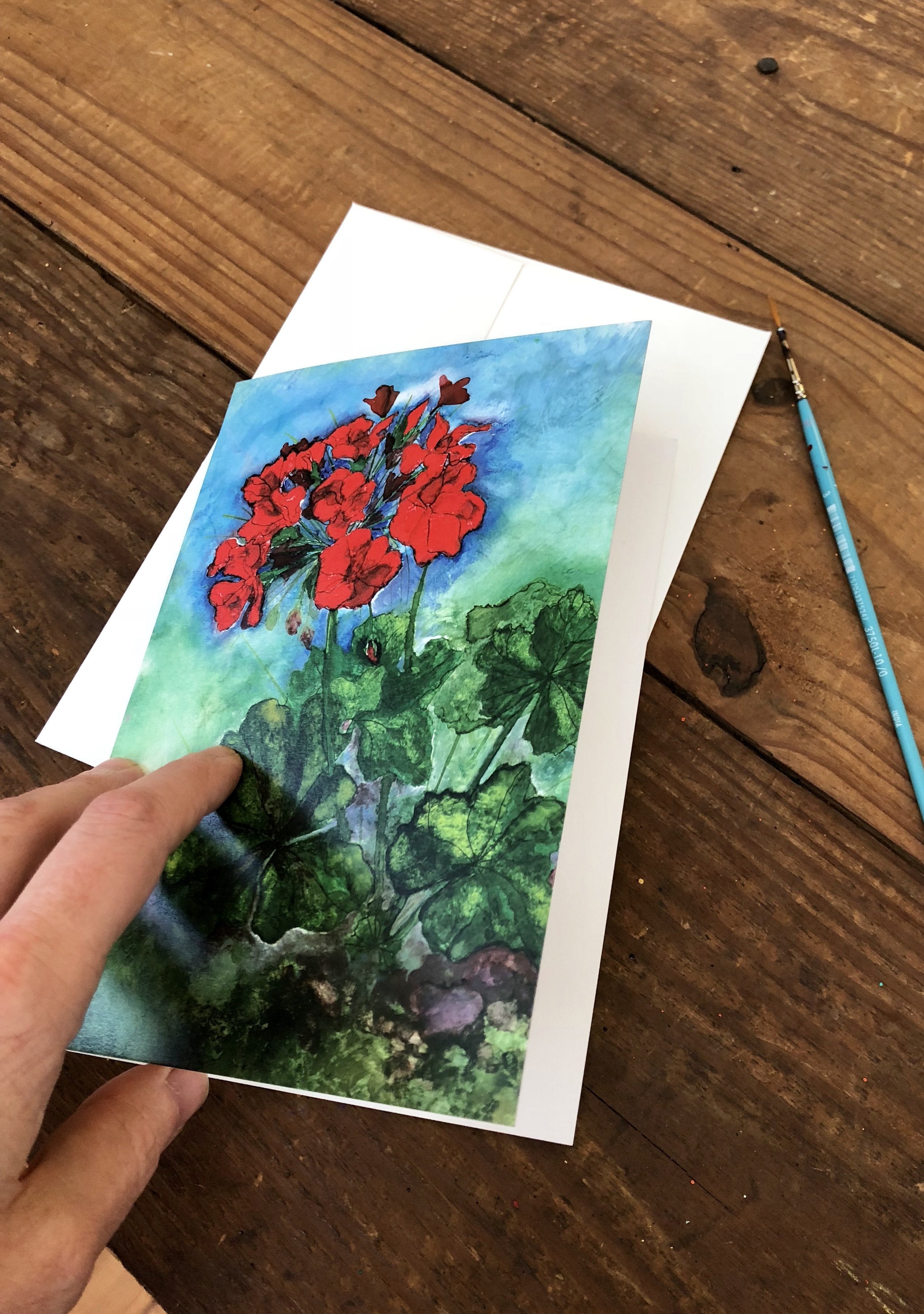 A vibrant red geranium greeting card featuring a mixed media painting with pen, oil paints, and alcohol inks, displayed with a white envelope.