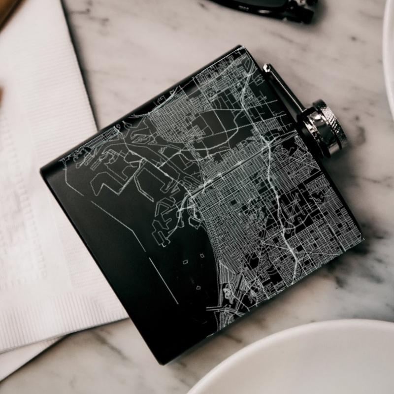 Matte black hip flask featuring a custom engraved map of Long Beach, California with coordinates, accompanied by a canvas bag and funnel.