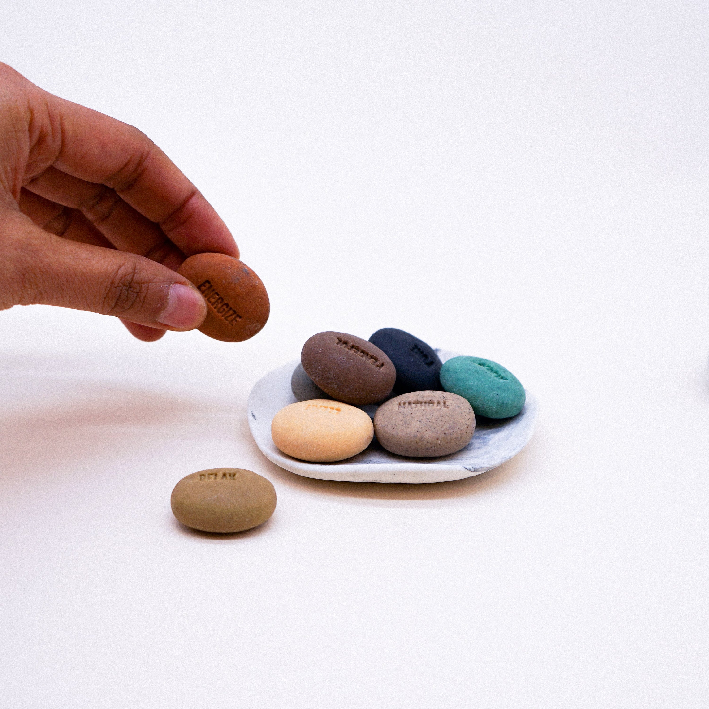 A set of handmade mood soaps in pebble design with a porcelain dish featuring a dark grey marble effect, showcasing assorted colors and mood words.