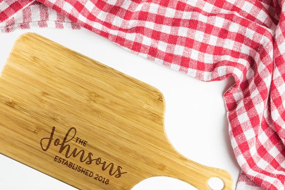 A personalized bamboo chopping board with a laser-etched design, showcasing its dual-purpose sides for food preparation and display, featuring a handle for easy hanging.