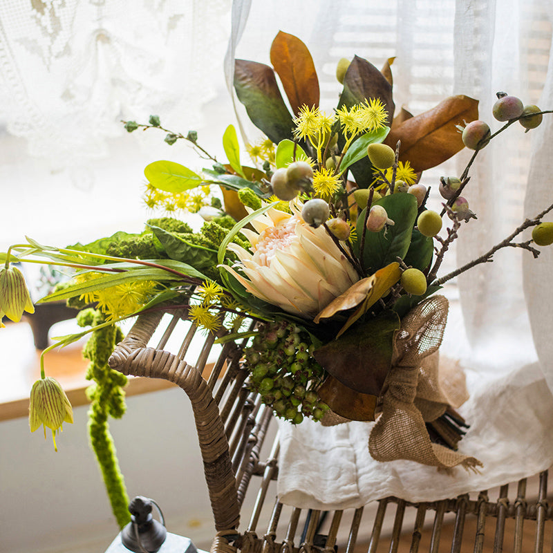 A 25-inch tall artificial bouquet featuring vibrant protea flowers, lush silk leaves, and eucalyptus seeds, arranged in a decorative style.