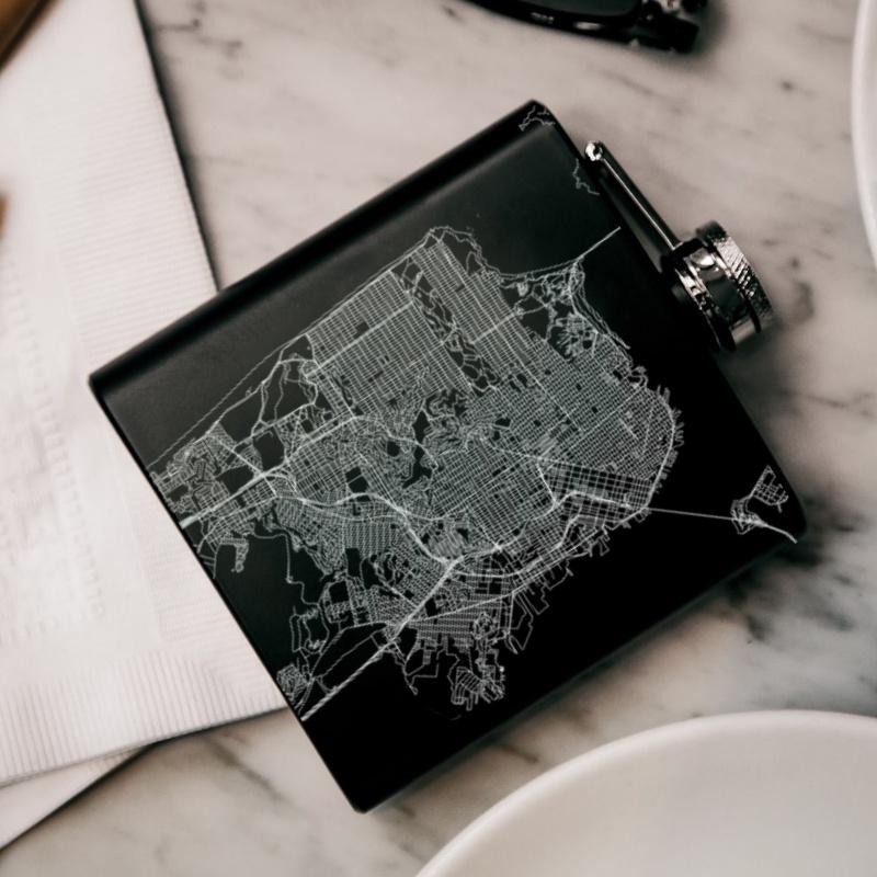 Matte black hip flask featuring a custom engraved map of San Francisco with coordinates, accompanied by a canvas bag and funnel.