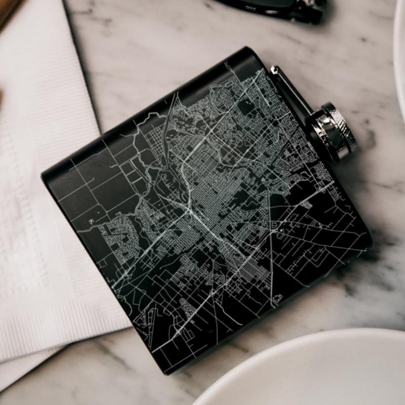 Matte black hip flask featuring a custom engraved map of Stockton, California with coordinates, accompanied by a canvas bag and funnel.