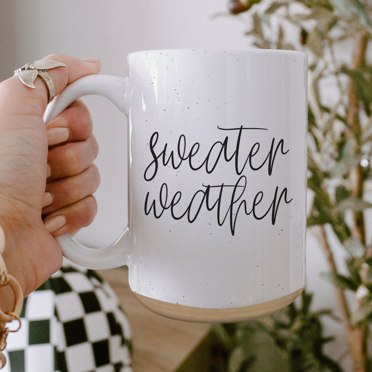 A cozy off-white ceramic mug with dark speckles, featuring a stylish black graphic that reads 'Sweater Weather', perfect for warm beverages.