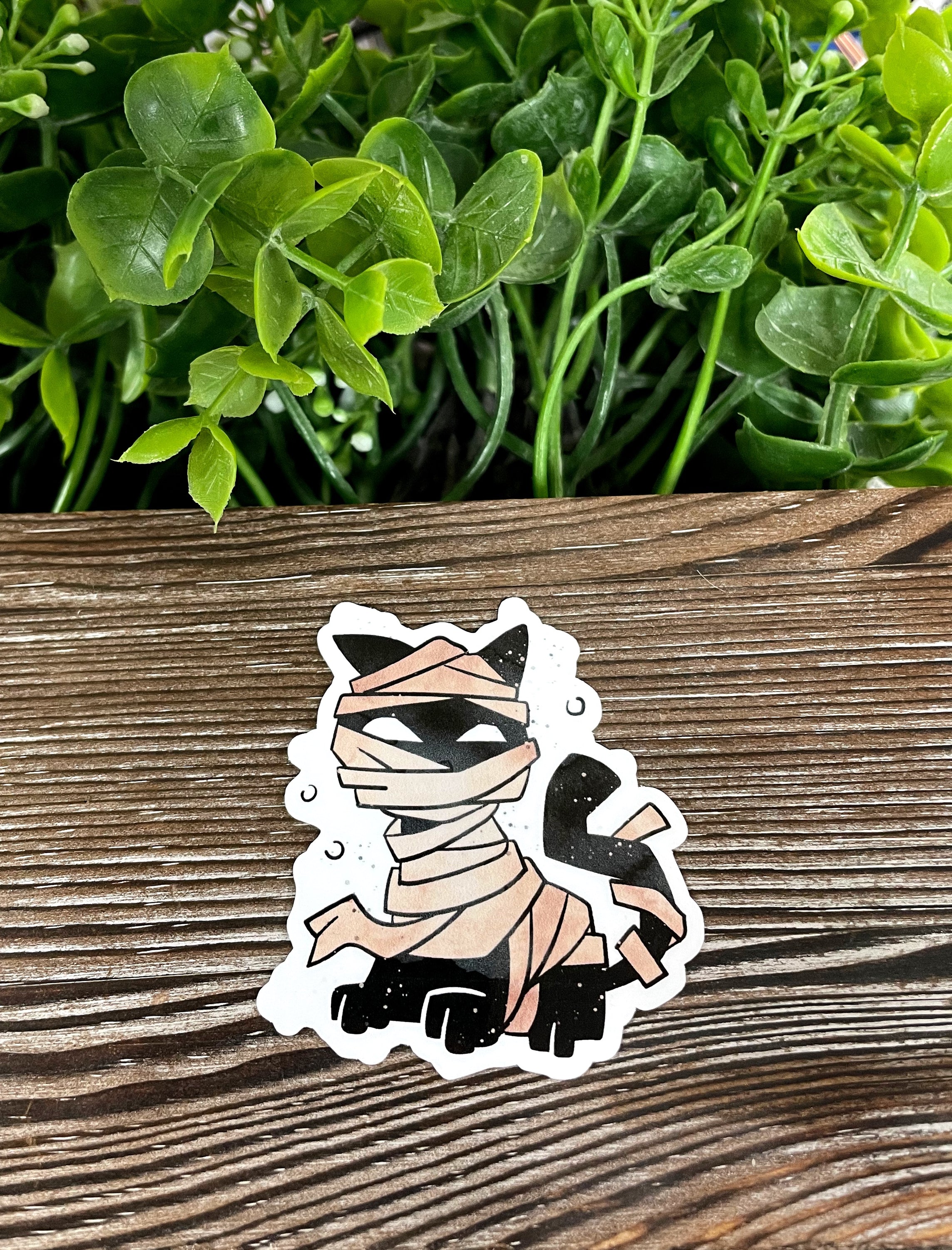 A die cut vinyl sticker featuring a black cat dressed in a mummy costume, showcasing vibrant colors and intricate details.