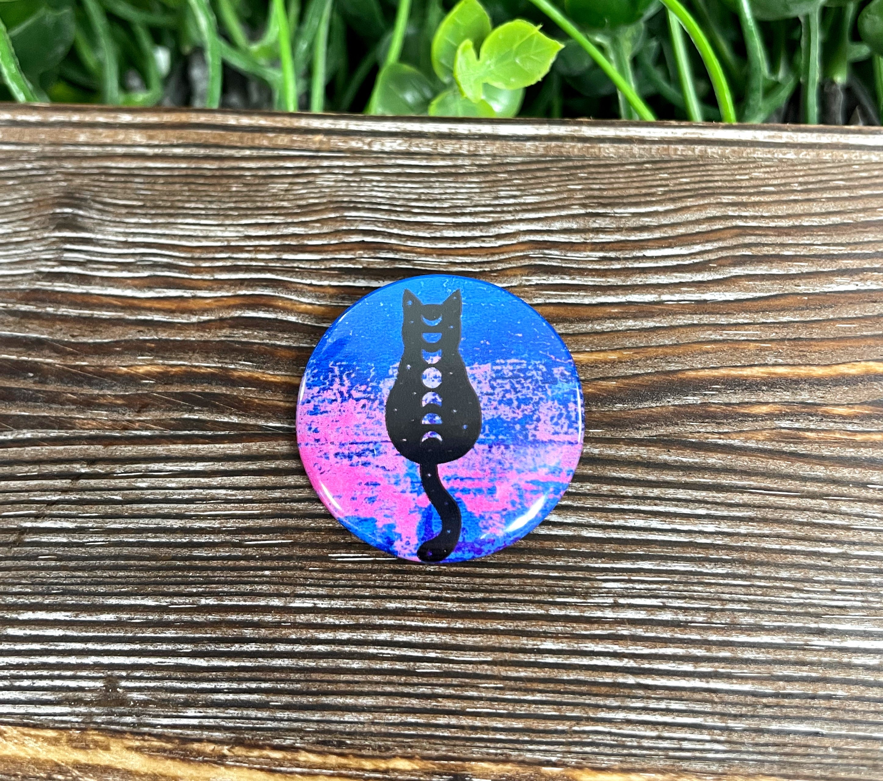 A 1.25-inch button featuring a black cat surrounded by moon phases in pink and blue colors, perfect for accessories.