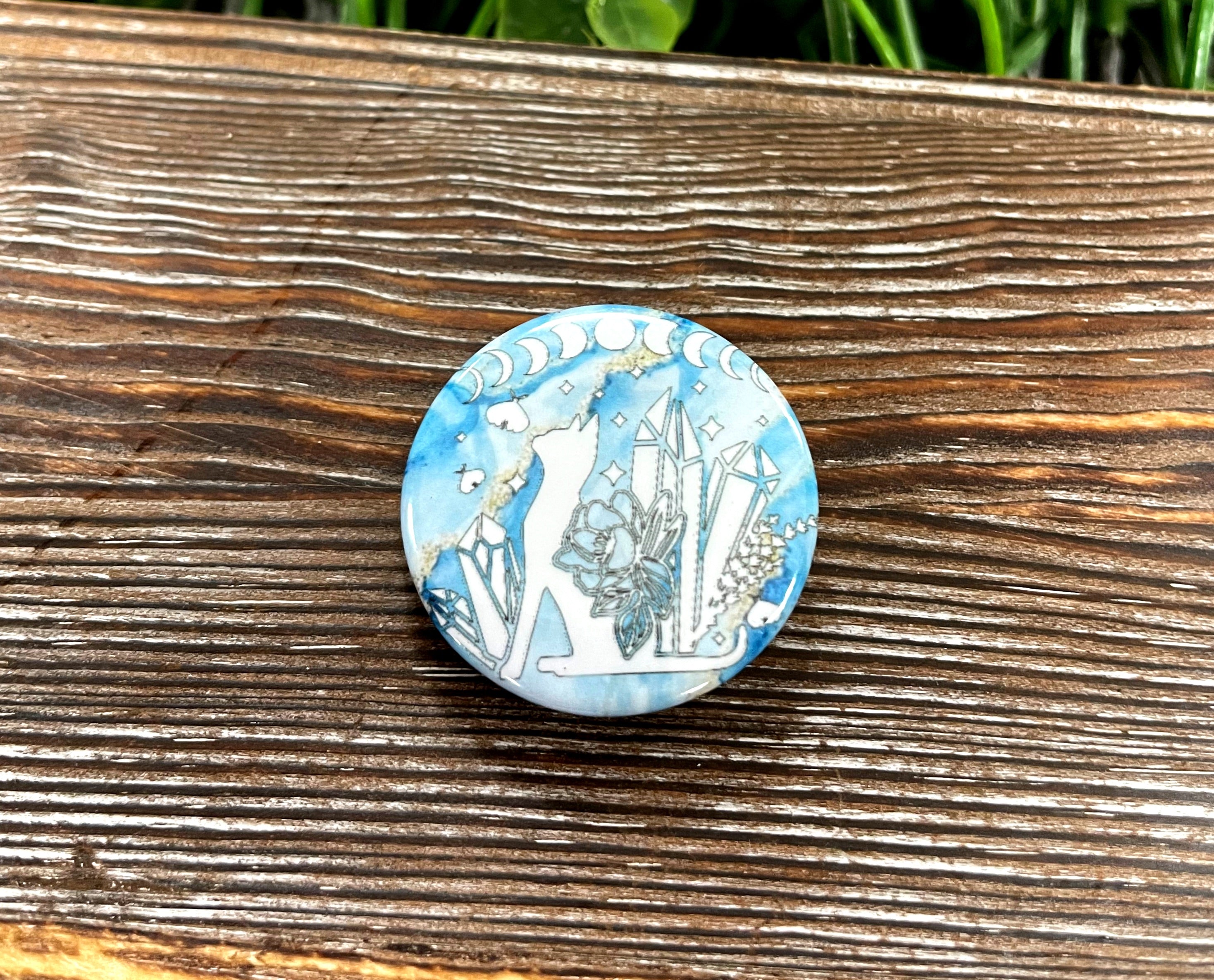 A 1.25-inch graphic art button featuring a blue marble cat design adorned with crystals, perfect for personalizing bags and accessories.