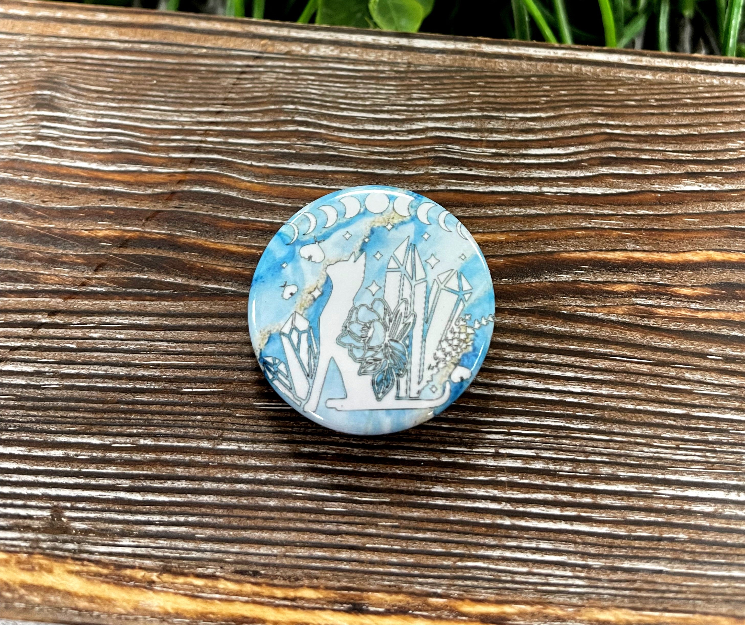 A 1.25-inch graphic art button featuring a blue marble cat design adorned with crystals, perfect for personalizing bags and accessories.