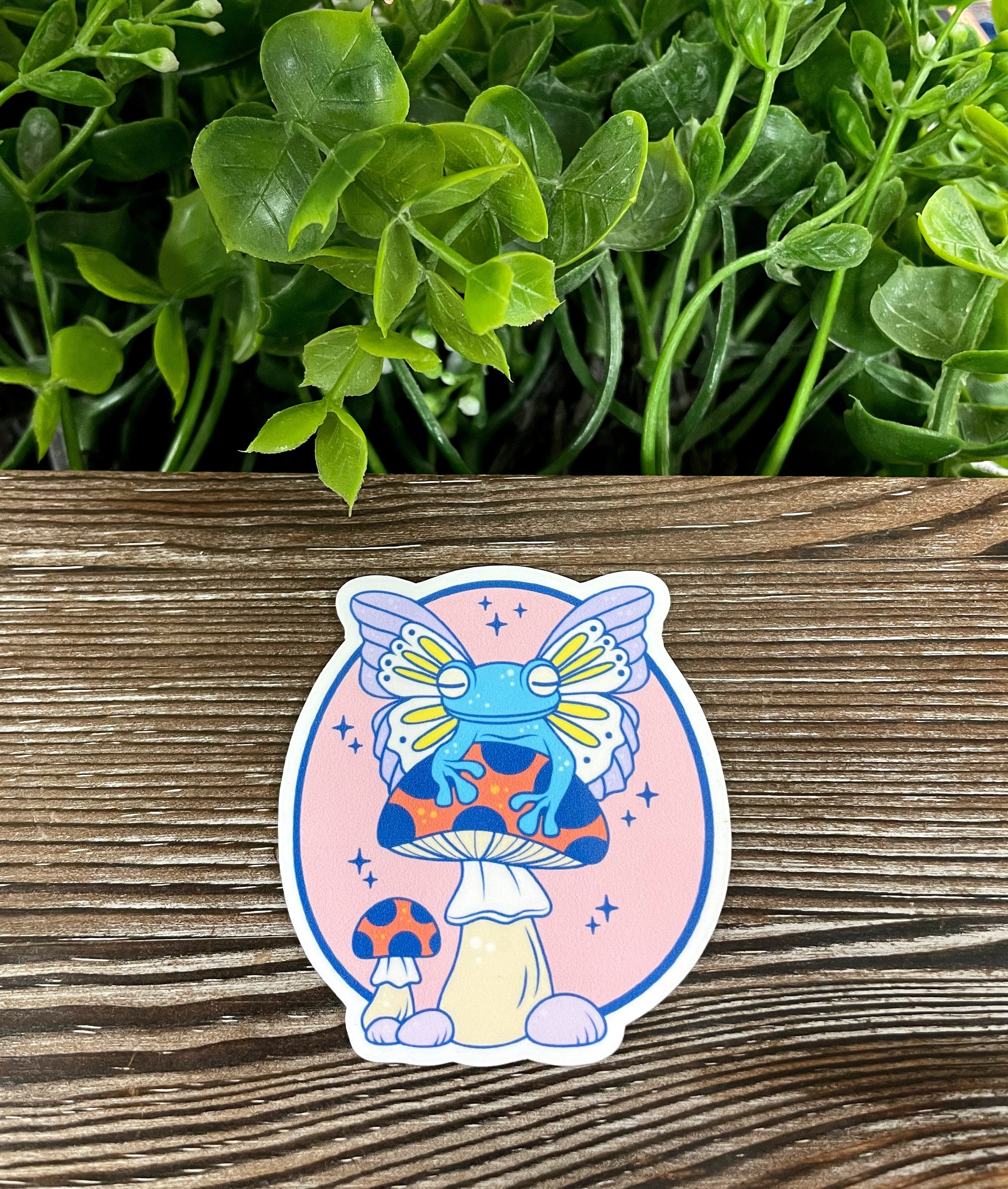 A whimsical die cut vinyl sticker featuring a frog with wings sitting on a mushroom, showcasing vibrant colors and intricate details.