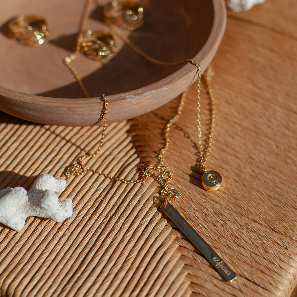 Layered Bar Bullet Necklace featuring recycled brass and bullet fragments, with a bar inscribed with 'peace' in Khmer, showcasing its unique design.