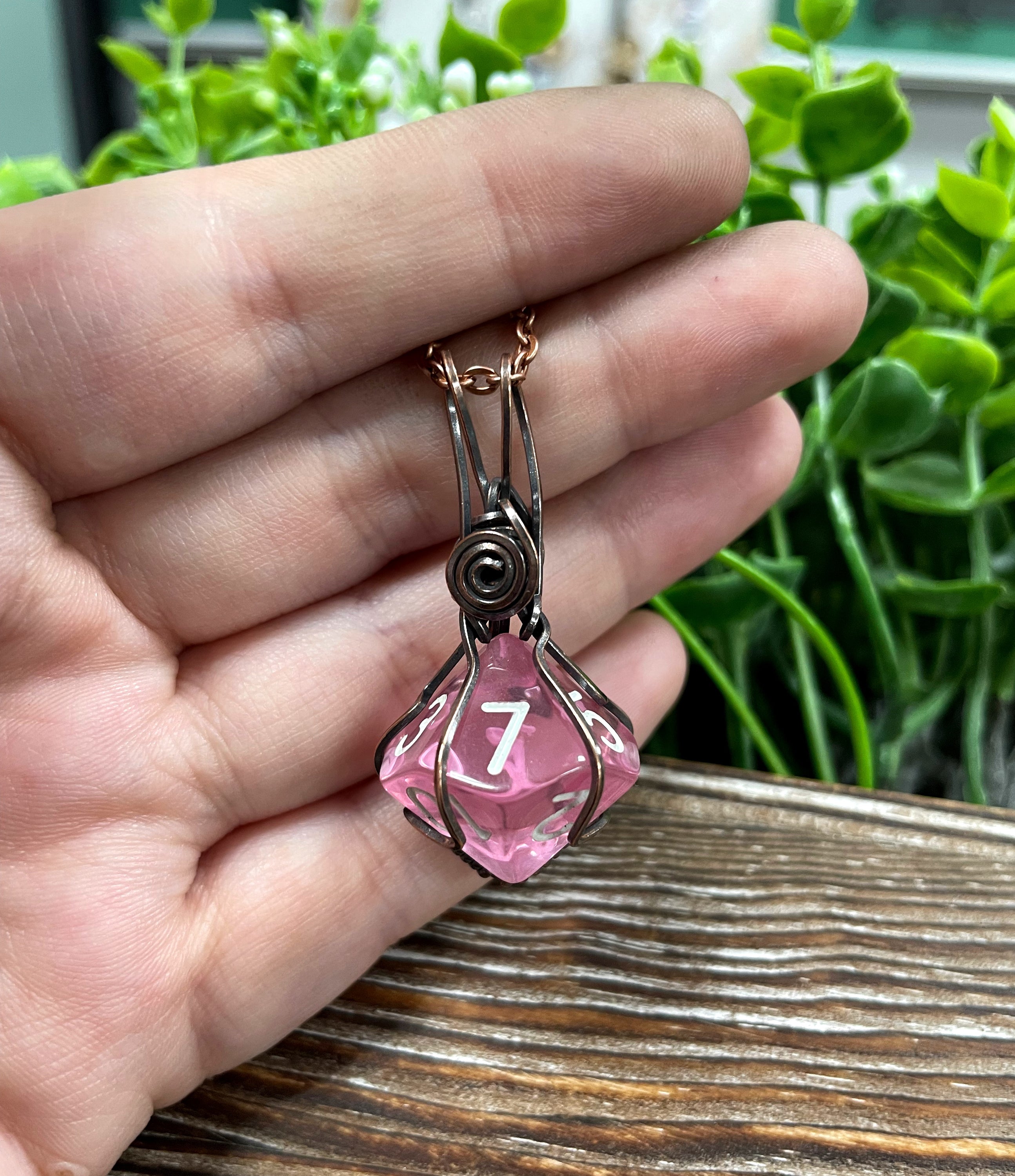 A beautiful pink D10 dice pendant wrapped in oxidized copper wire, hanging from a 21-inch suede leather cord, showcasing its unique design.