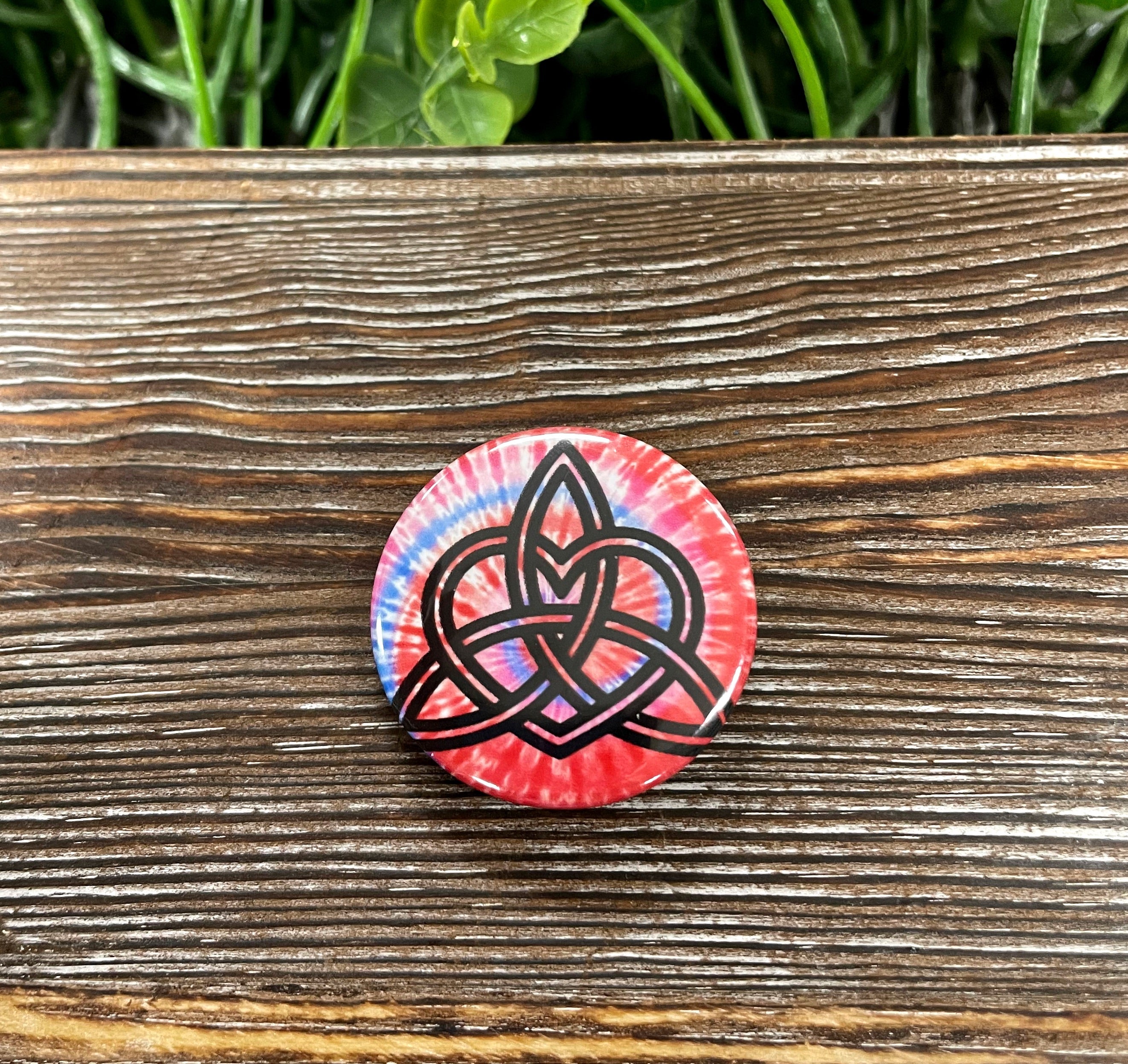 A vibrant 1.25-inch button featuring a red and blue tie dye pattern with a Triquetra graphic design, perfect for accessories.