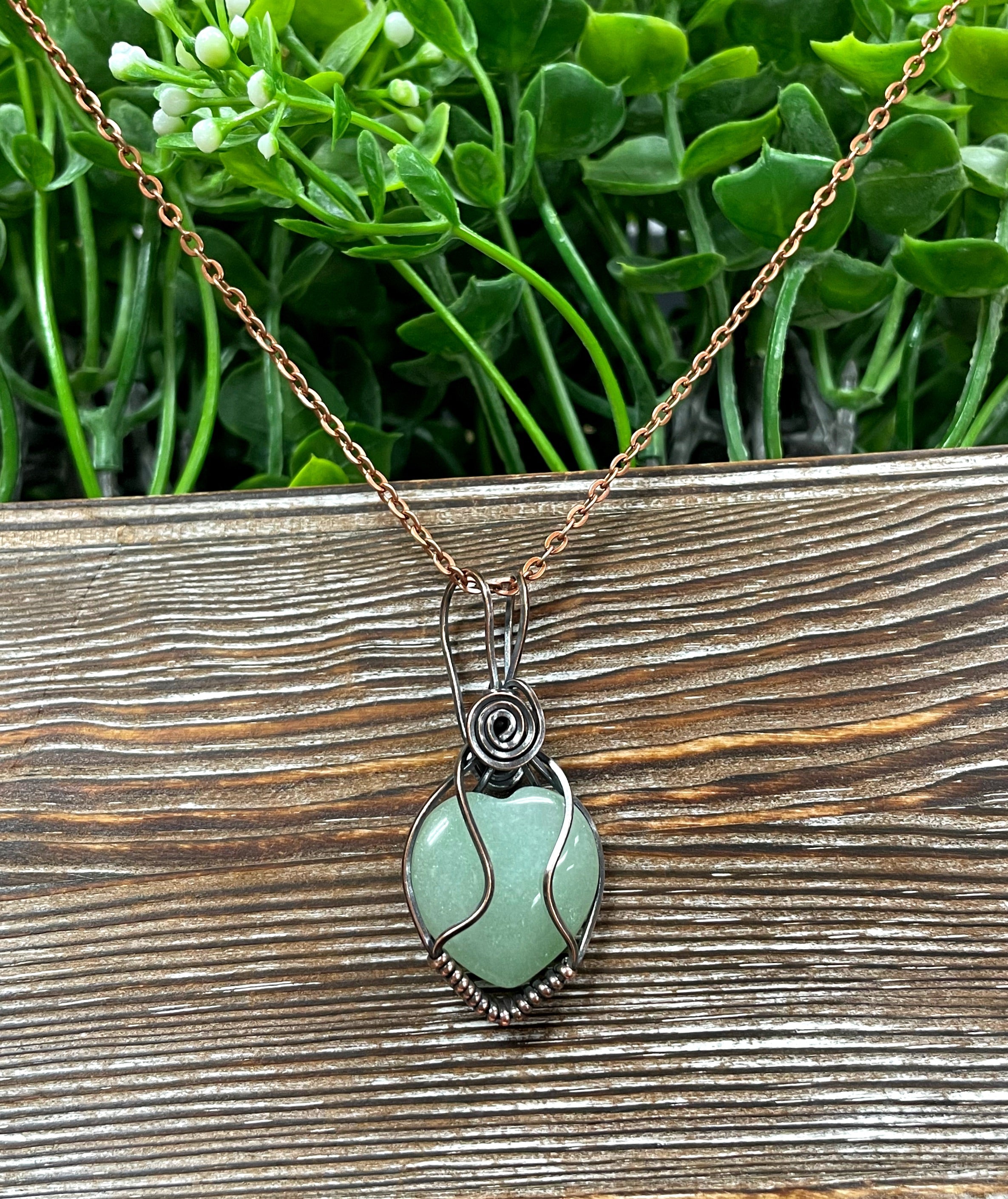 Small heart-shaped green aventurine gemstone pendant wrapped in oxidized copper wire, displayed on a 21-inch cord.