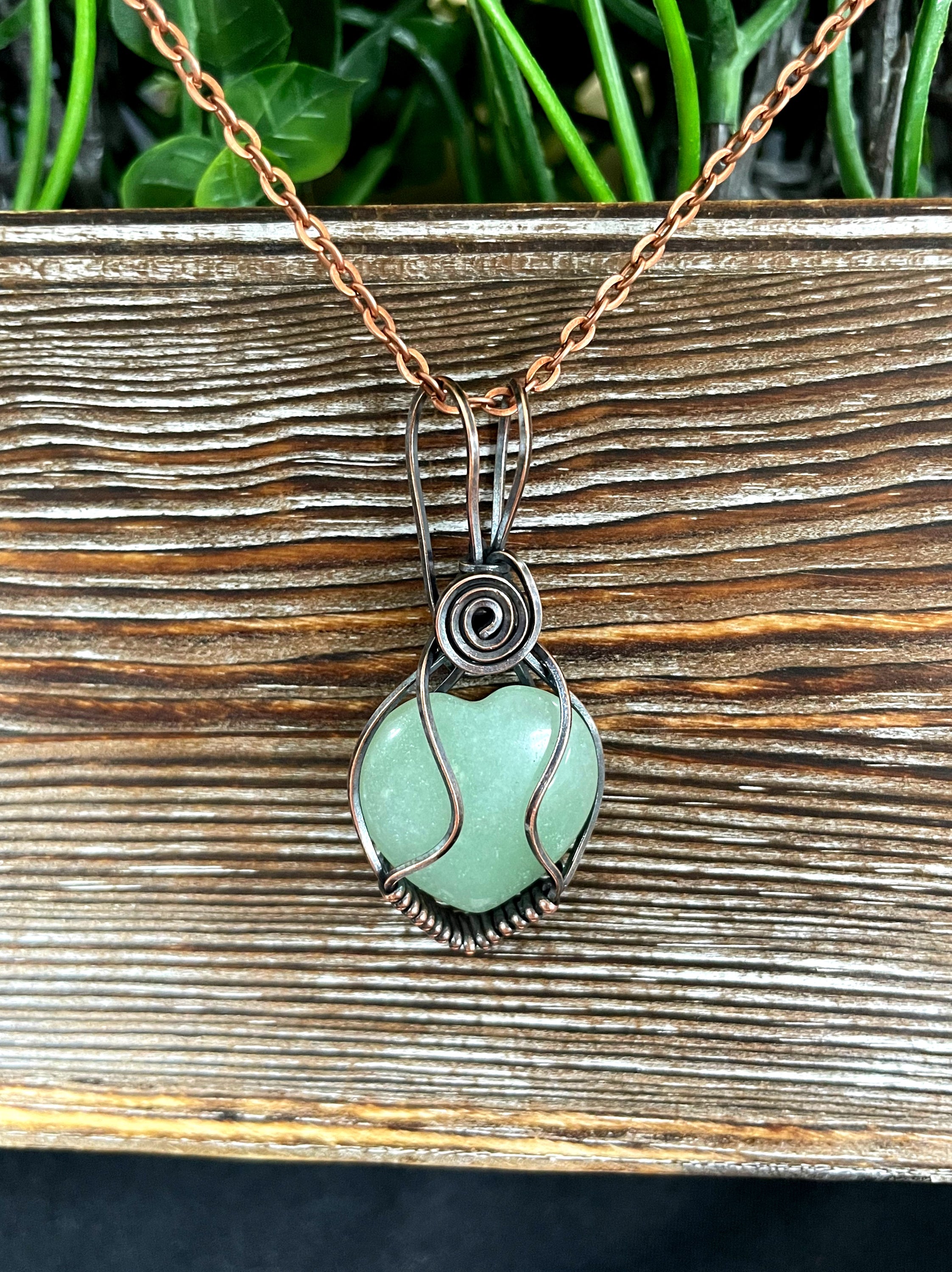 Small heart-shaped green aventurine gemstone pendant wrapped in oxidized copper wire, displayed on a 21-inch cord.