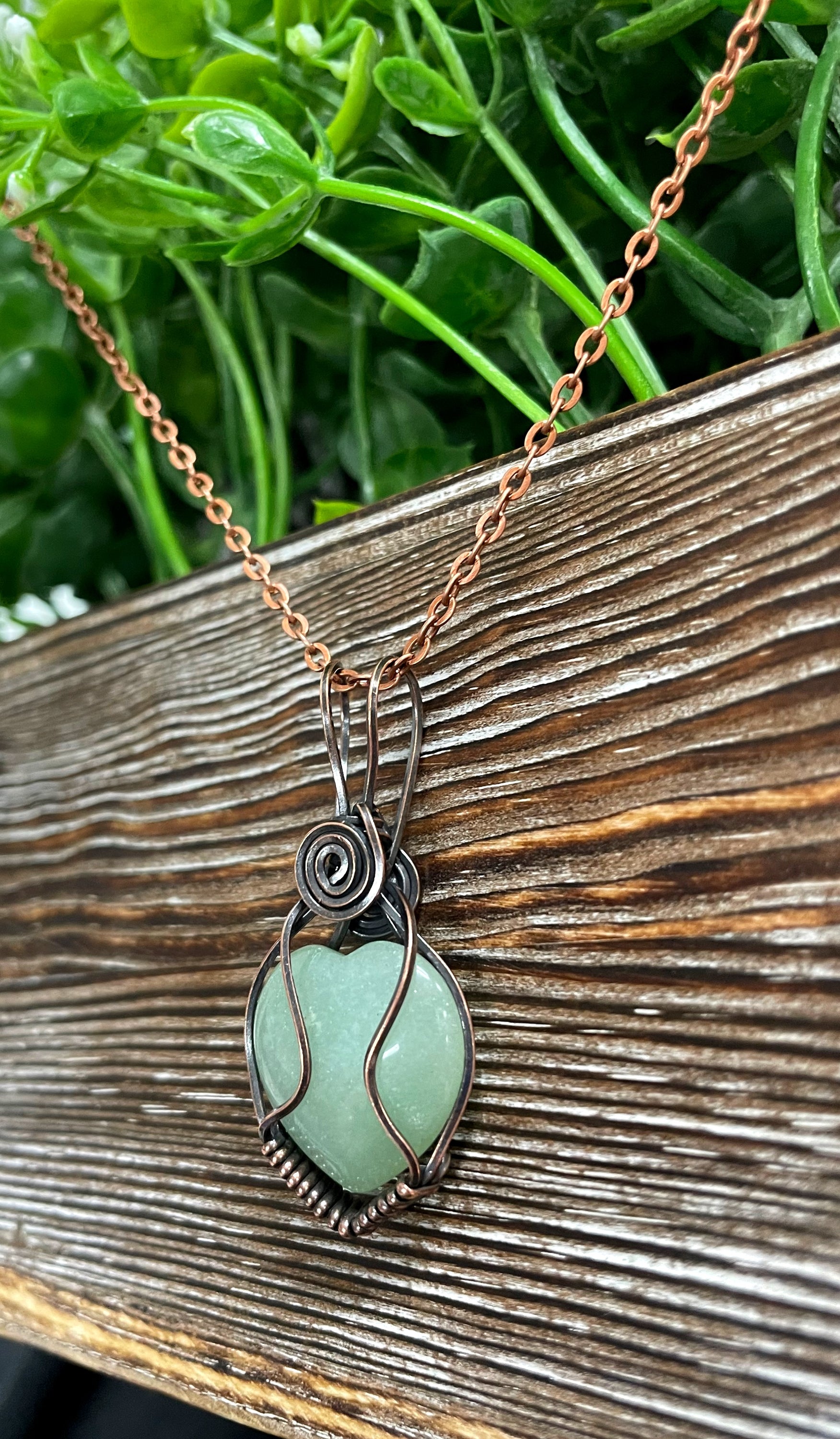 Small heart-shaped green aventurine gemstone pendant wrapped in oxidized copper wire, displayed on a 21-inch cord.