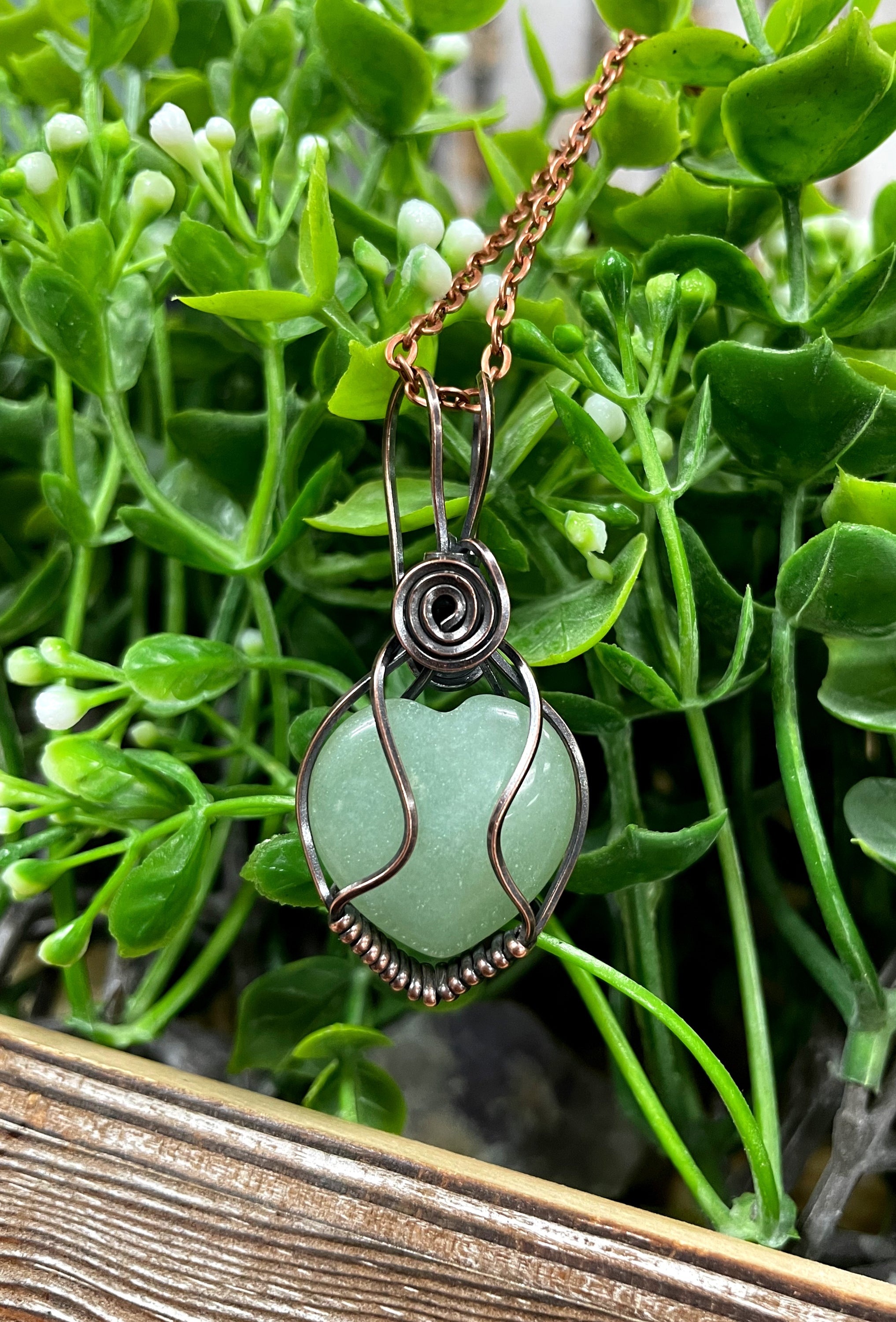 Small heart-shaped green aventurine gemstone pendant wrapped in oxidized copper wire, displayed on a 21-inch cord.