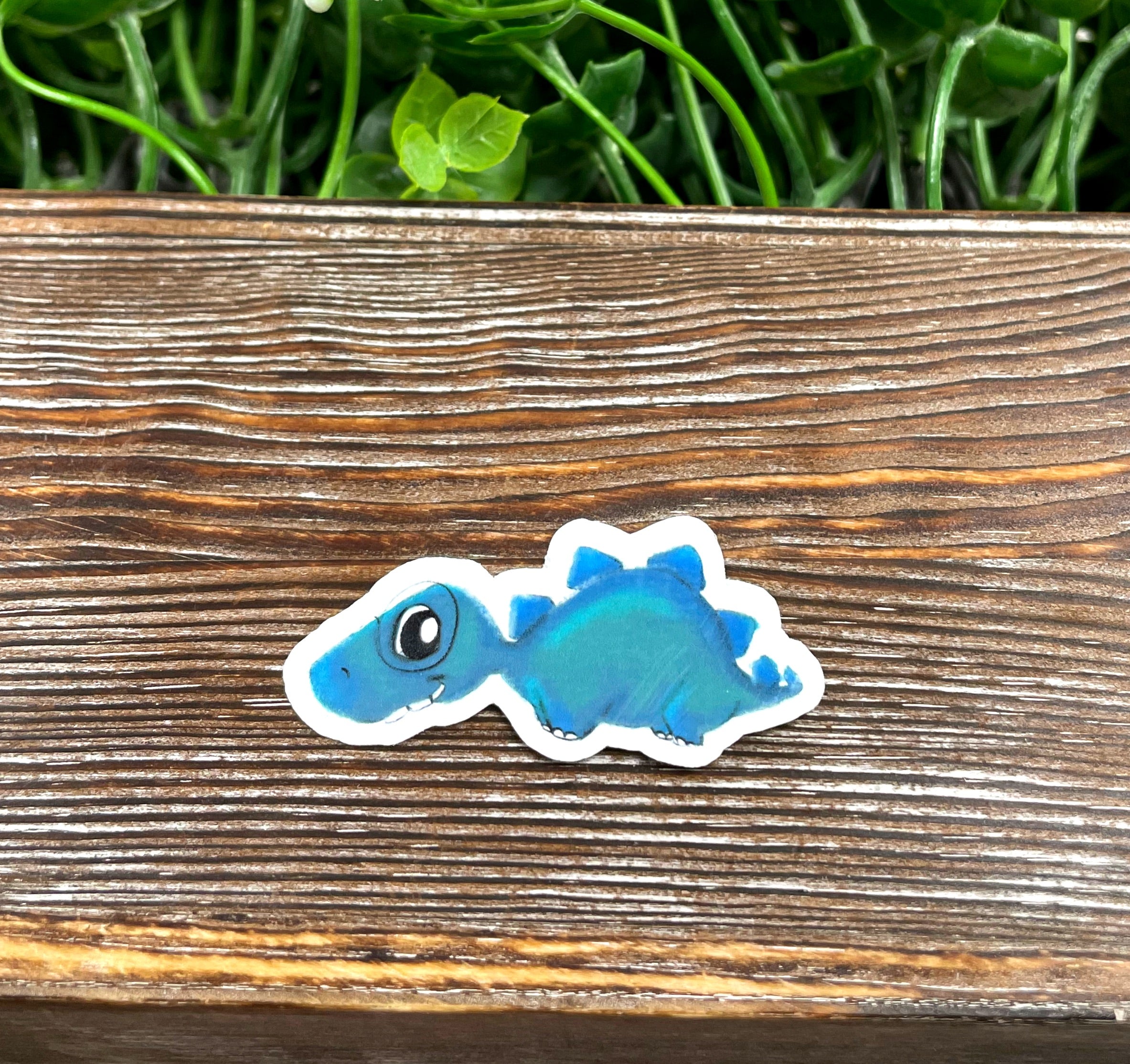 A vibrant watercolor blue dinosaur die cut sticker on a smooth surface, showcasing its intricate design and colors.