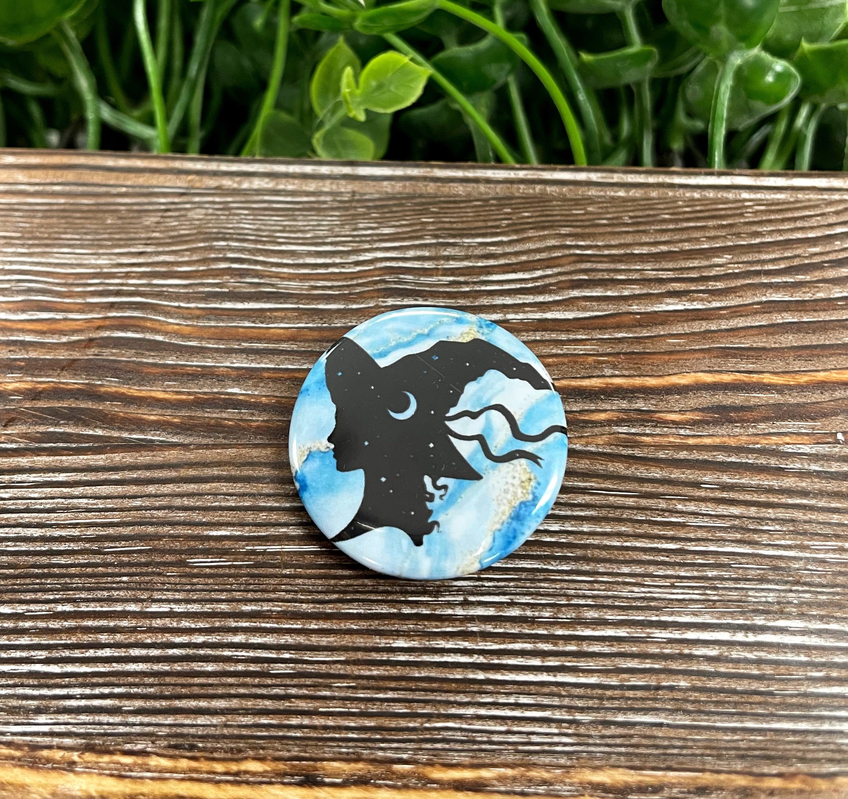 A 1.25-inch button featuring a witch silhouette against a moon and stars background, showcasing a whimsical and mystical design.