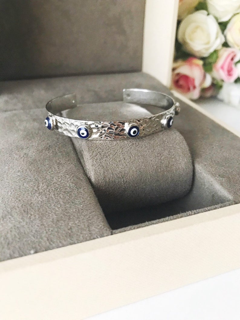 A stunning Evil Eye Bangle Bracelet featuring a movable oval charm, available in rose gold and silver finishes with customizable bead colors.