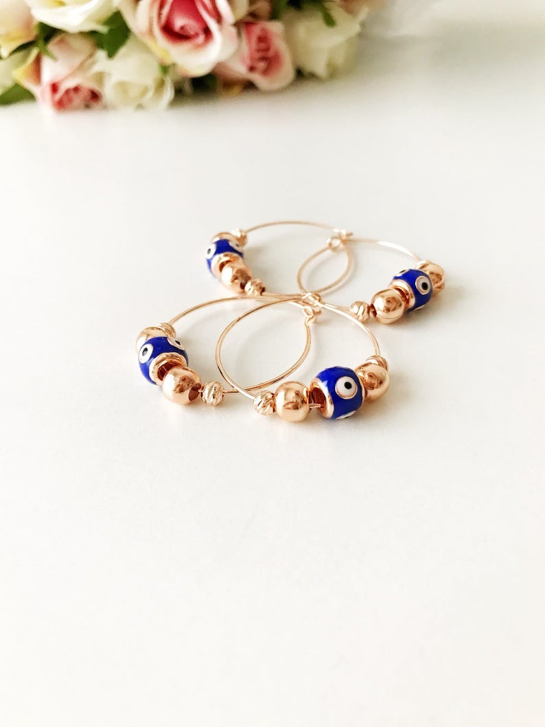 A pair of blue evil eye hoop earrings with a rose gold finish, featuring a small blue charm and a minimalist design.