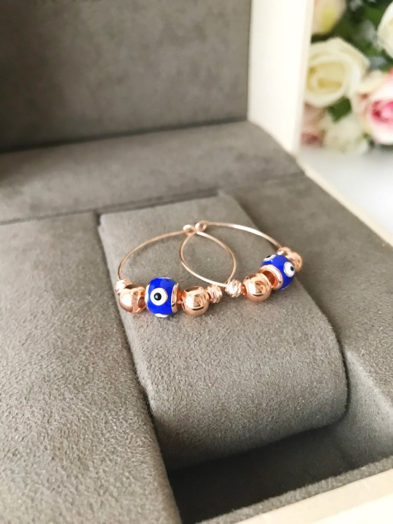 A pair of blue evil eye hoop earrings with a rose gold finish, featuring a small blue charm and a minimalist design.