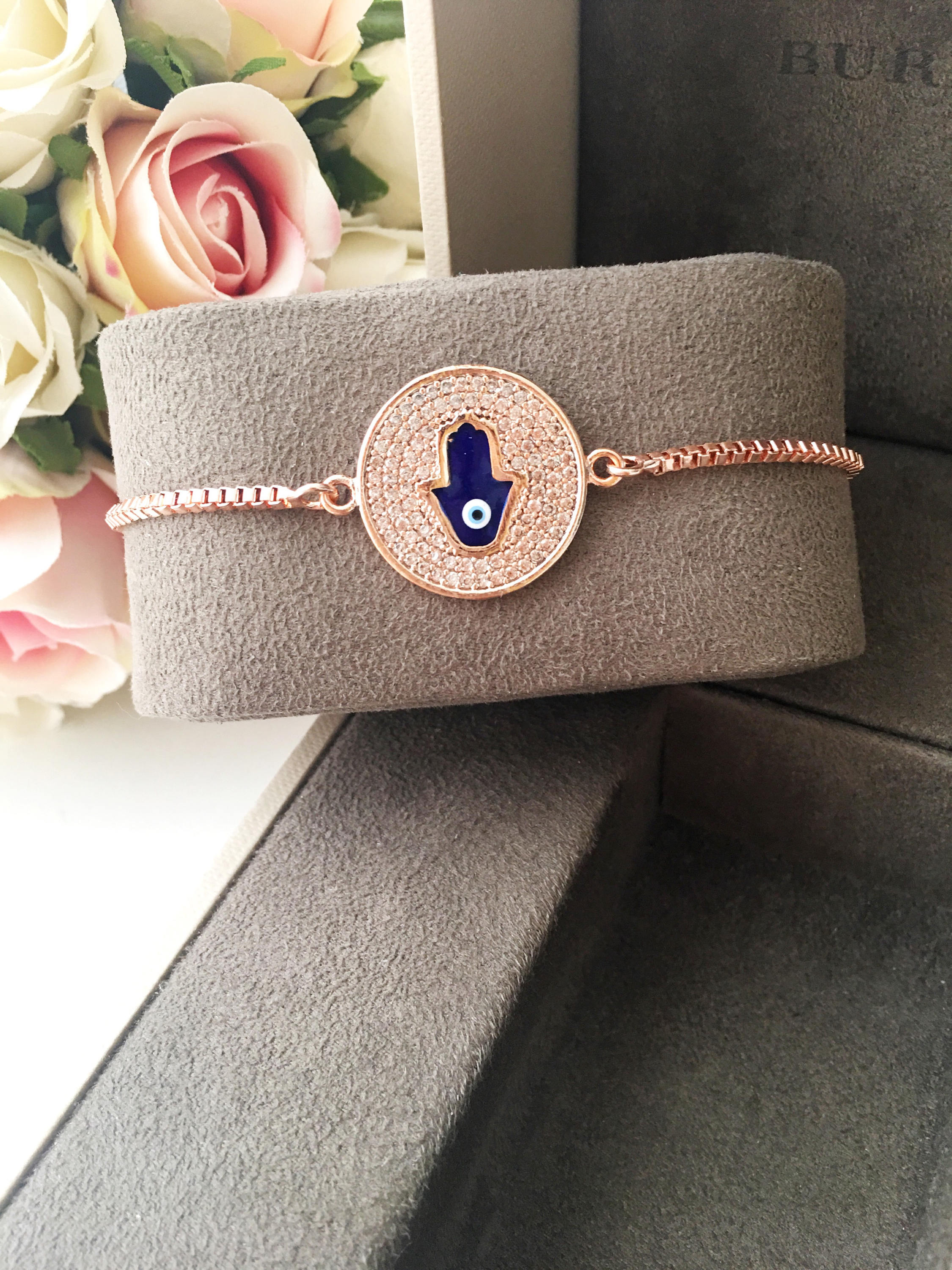 A beautiful Blue Hamsa Evil Eye Bracelet featuring a blue evil eye charm and a rose gold stainless steel disc, adjustable for a perfect fit.