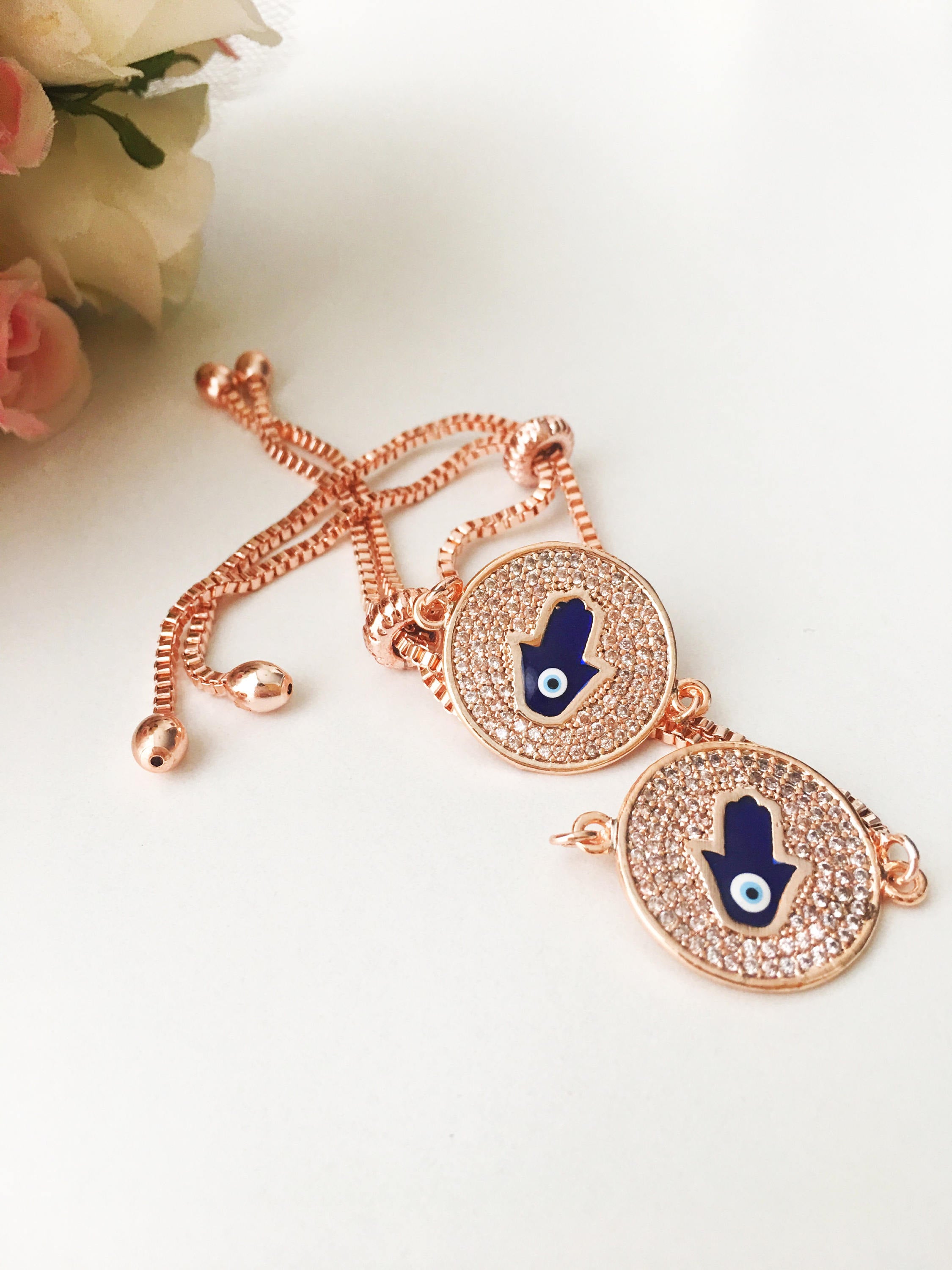 A beautiful Blue Hamsa Evil Eye Bracelet featuring a blue evil eye charm and a rose gold stainless steel disc, adjustable for a perfect fit.