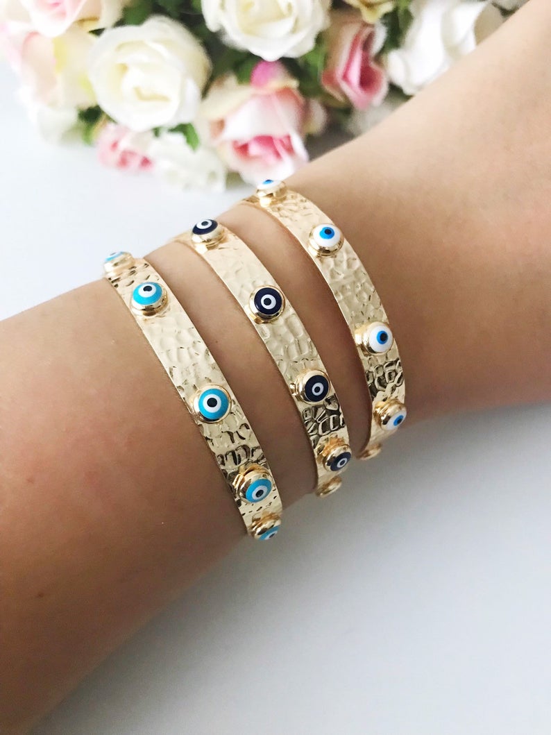 A beautiful Evil Eye Bracelet featuring seed beads and a gold finish, showcasing various styles including a turquoise cuff and blue oval bangle.