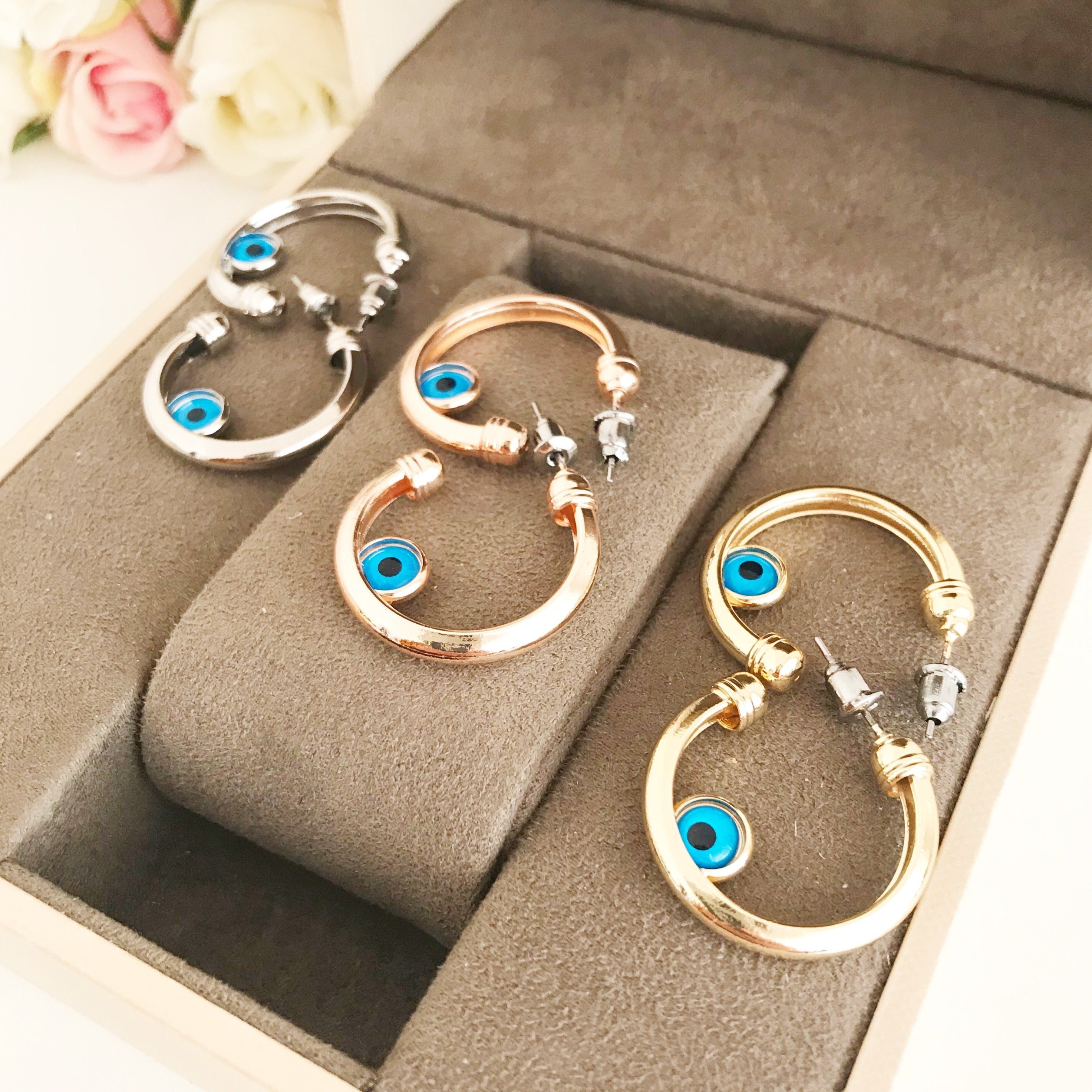 A pair of handmade evil eye hoop earrings featuring a blue evil eye charm, available in rose gold, gold, and silver finishes.