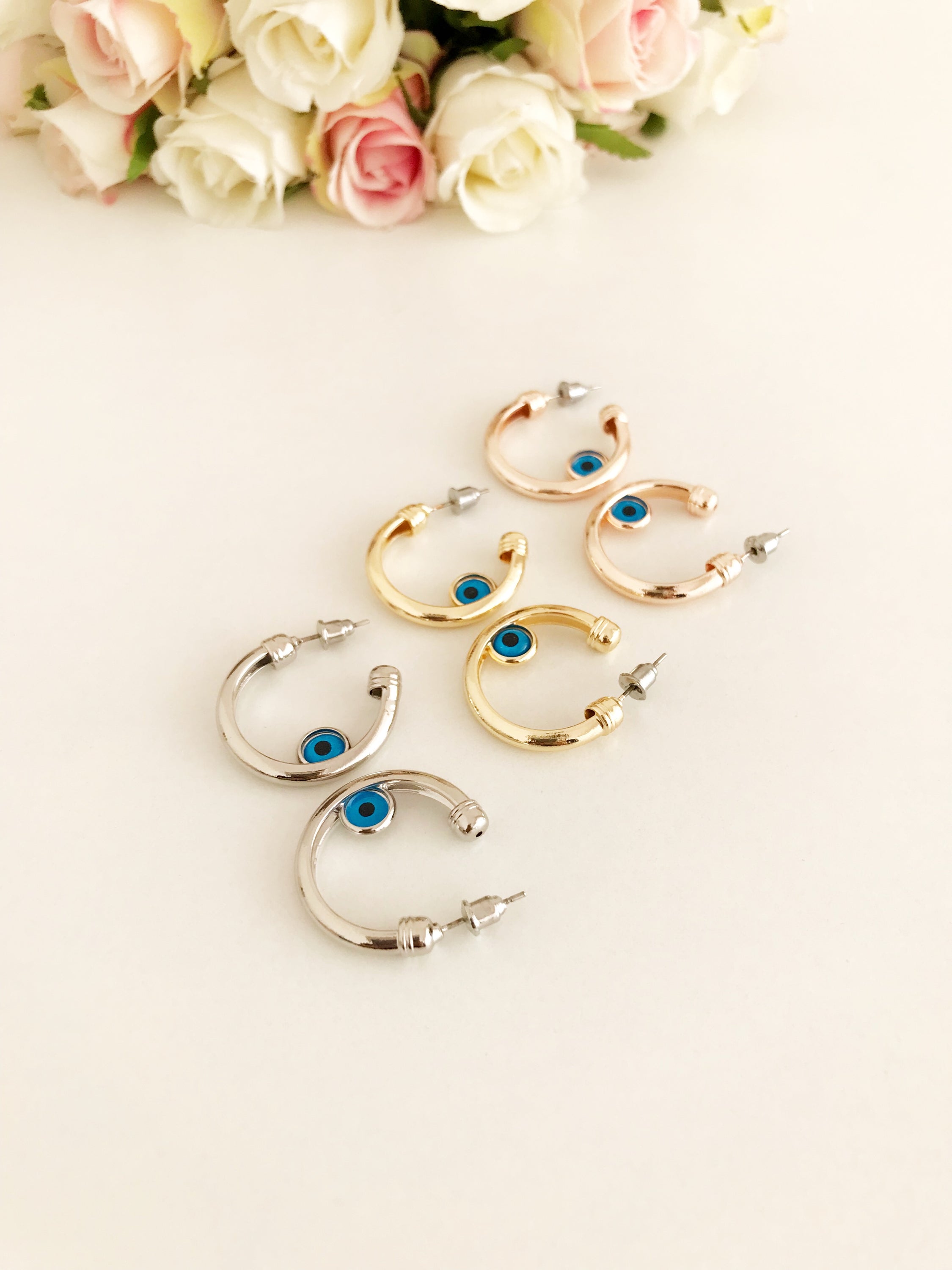 A pair of handmade evil eye hoop earrings featuring a blue evil eye charm, available in rose gold, gold, and silver finishes.