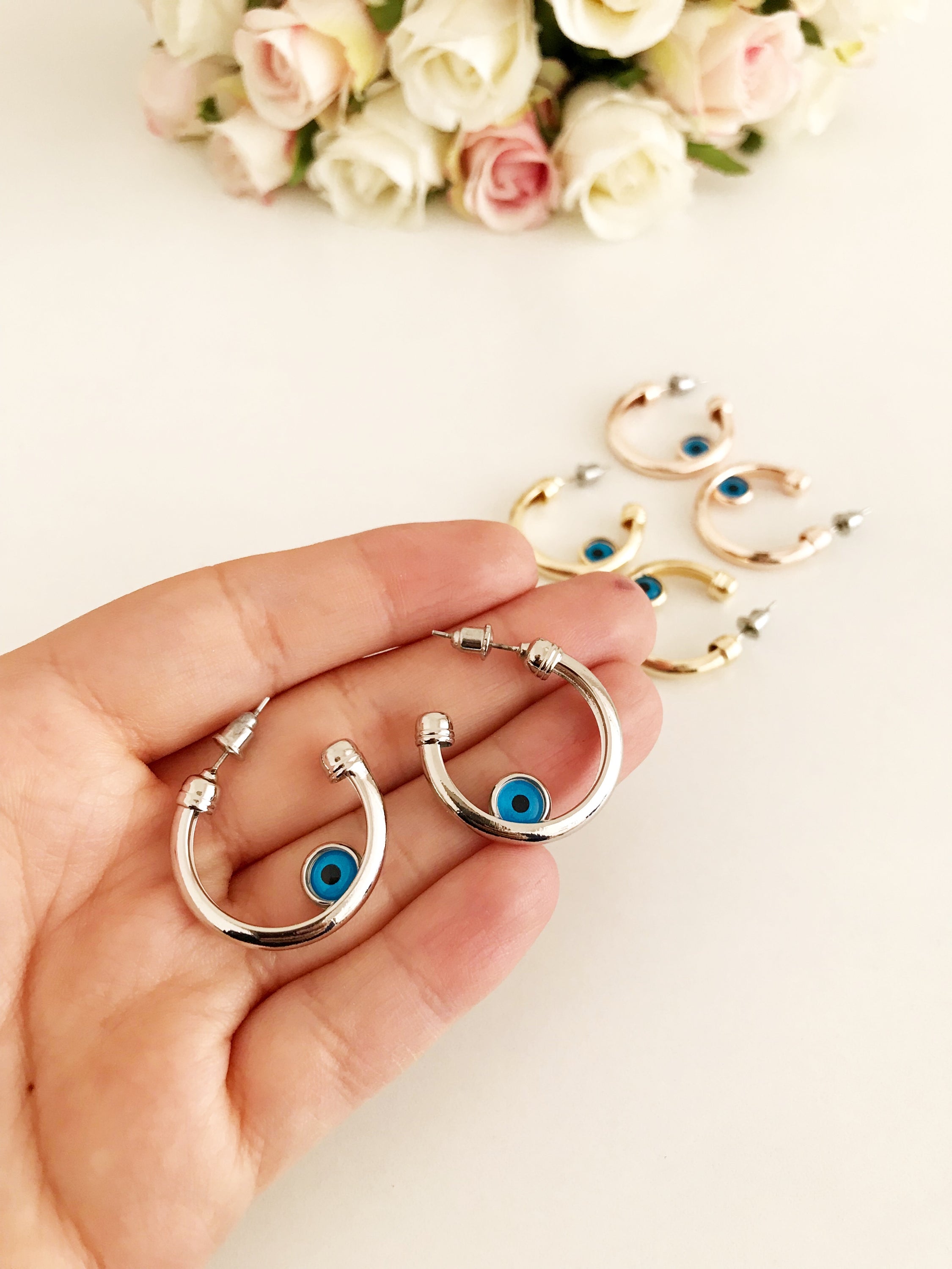 A pair of handmade evil eye hoop earrings featuring a blue evil eye charm, available in rose gold, gold, and silver finishes.